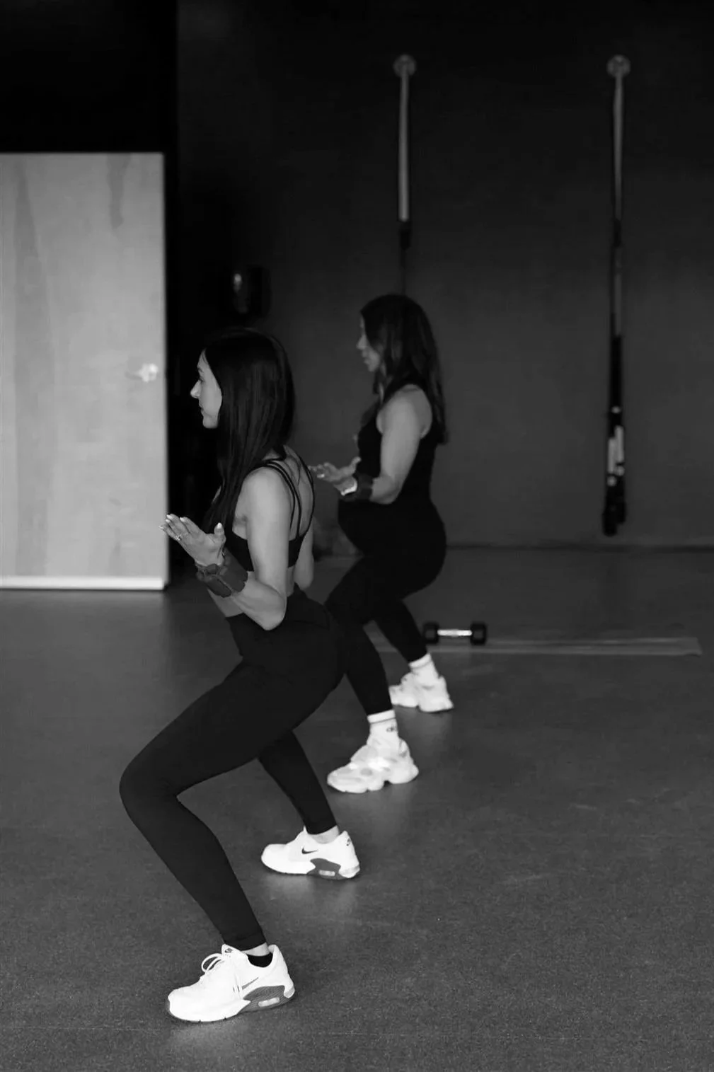 Two women exercising in a gym, doing squats with their hands pressed together in front of their chests, wearing workout clothes and sneakers.