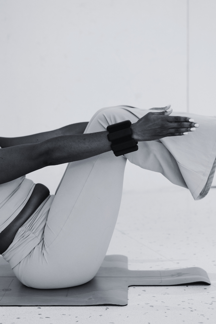 A person performing a stretching exercise on a yoga mat, with one arm extended and hand resting on their knee, black smartwatch on wrist, in a minimalistic indoor setting.