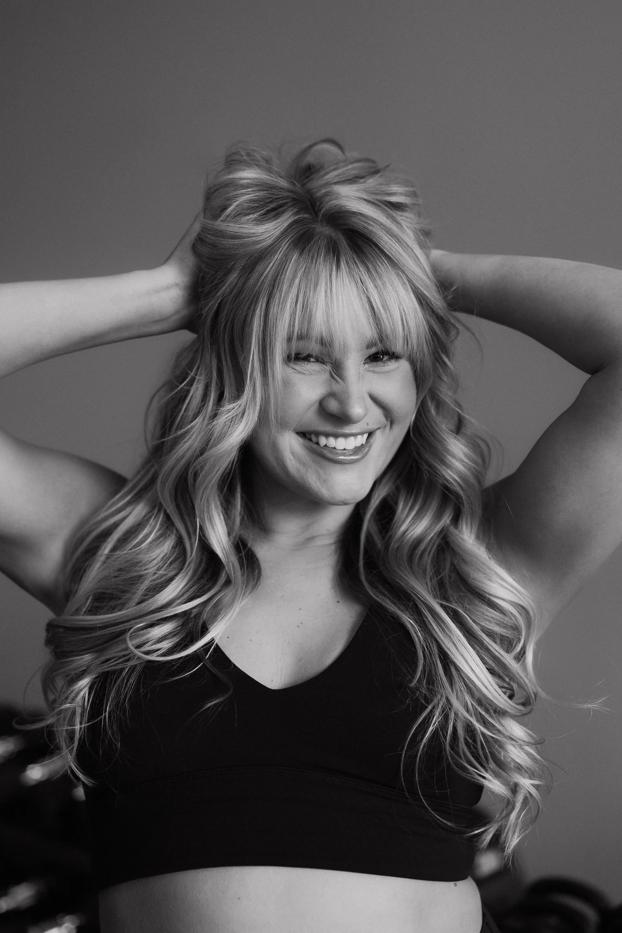 A woman with long, wavy blonde hair smiling, with her hands in her hair, wearing a black sports bra.
