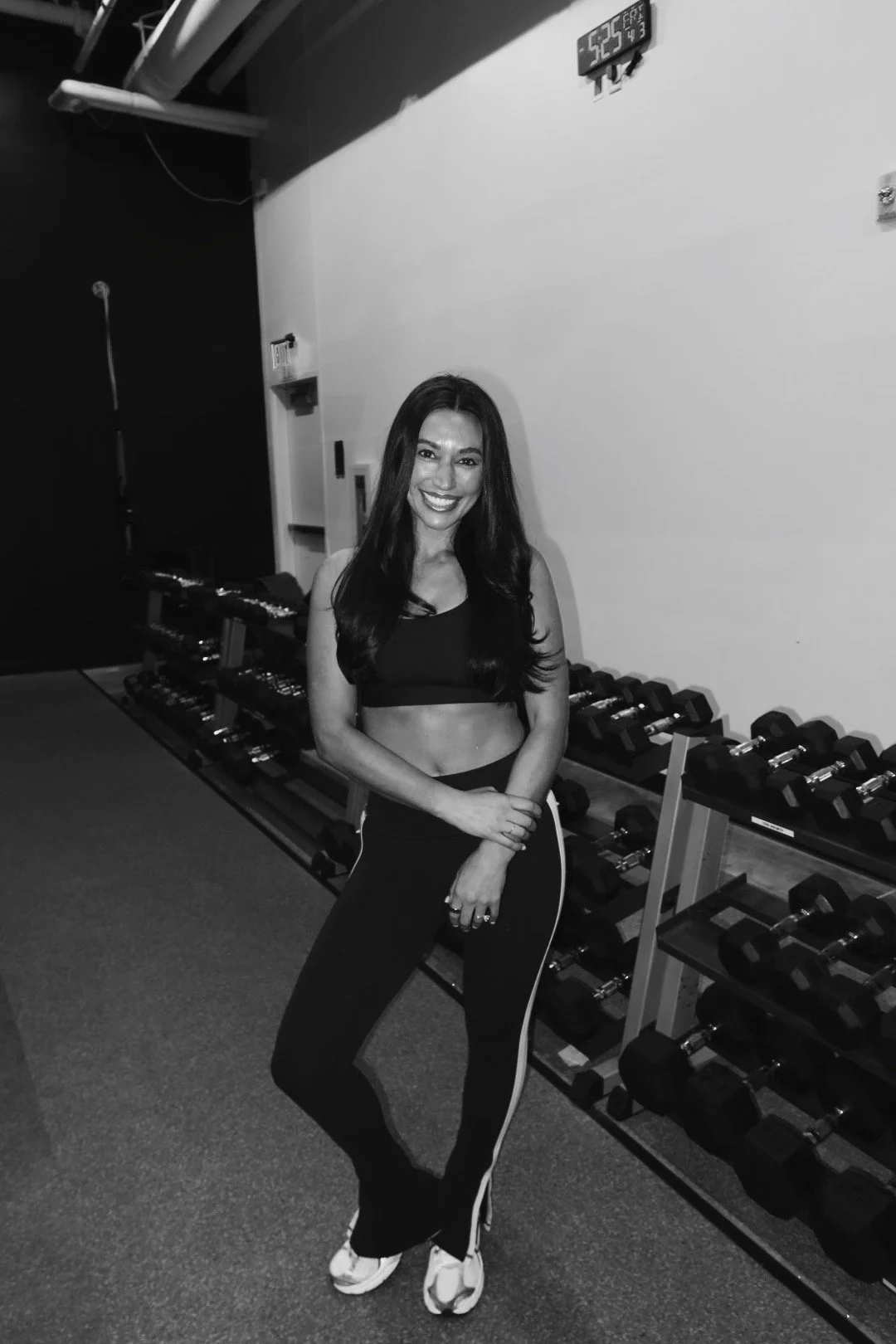 A smiling woman with long dark hair in workout attire standing in a gym, with exercise equipment in the background.