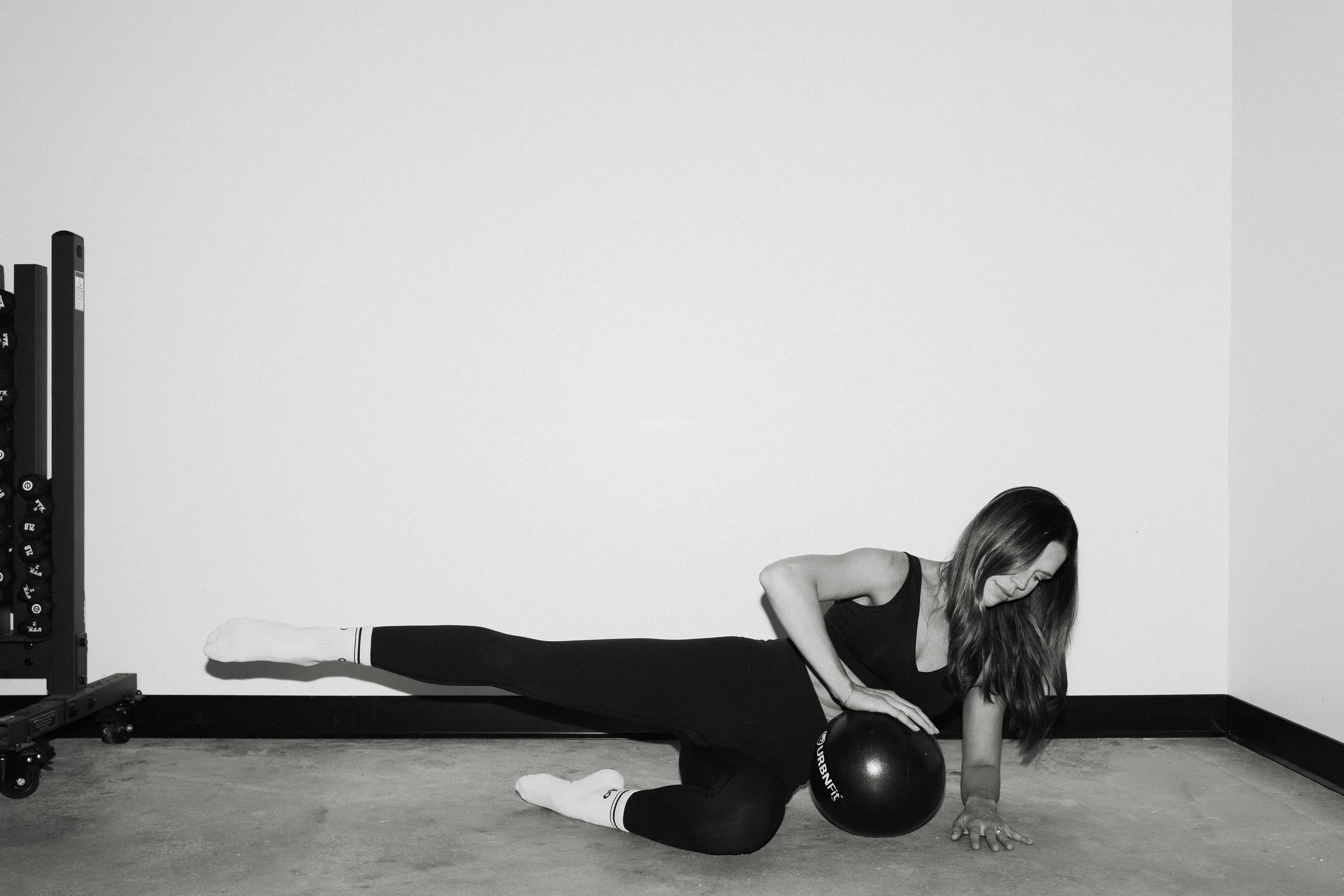 Two women exercising in a gym, squatting while facing a wall, with fitness equipment in the background.