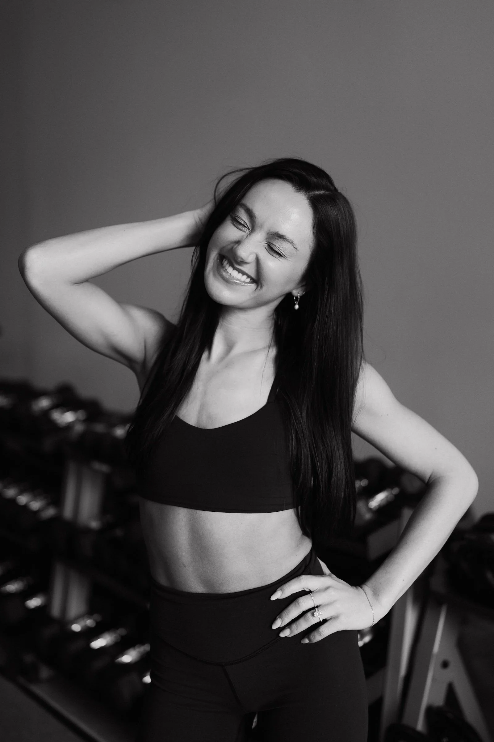 Black and white photograph of a woman with long dark hair, wearing a sports bra and high-waisted leggings, smiling with her eyes closed and her hand behind her head, standing in a gym or fitness studio.