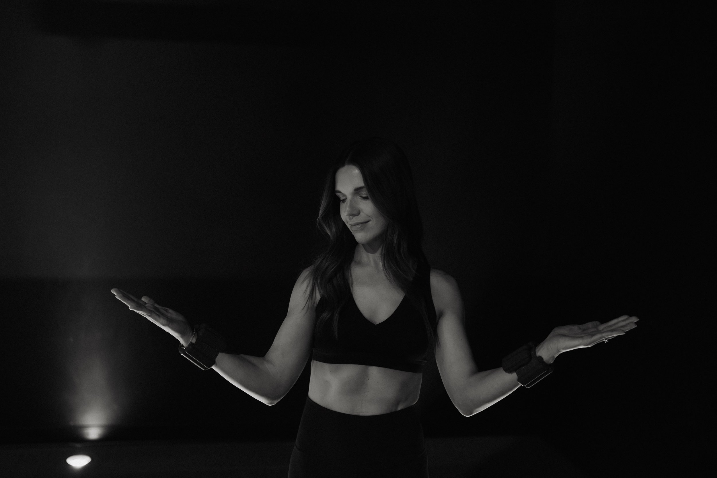 Two women exercising in a gym, doing squats with their hands pressed together in front of their chests, wearing workout clothes and sneakers.
