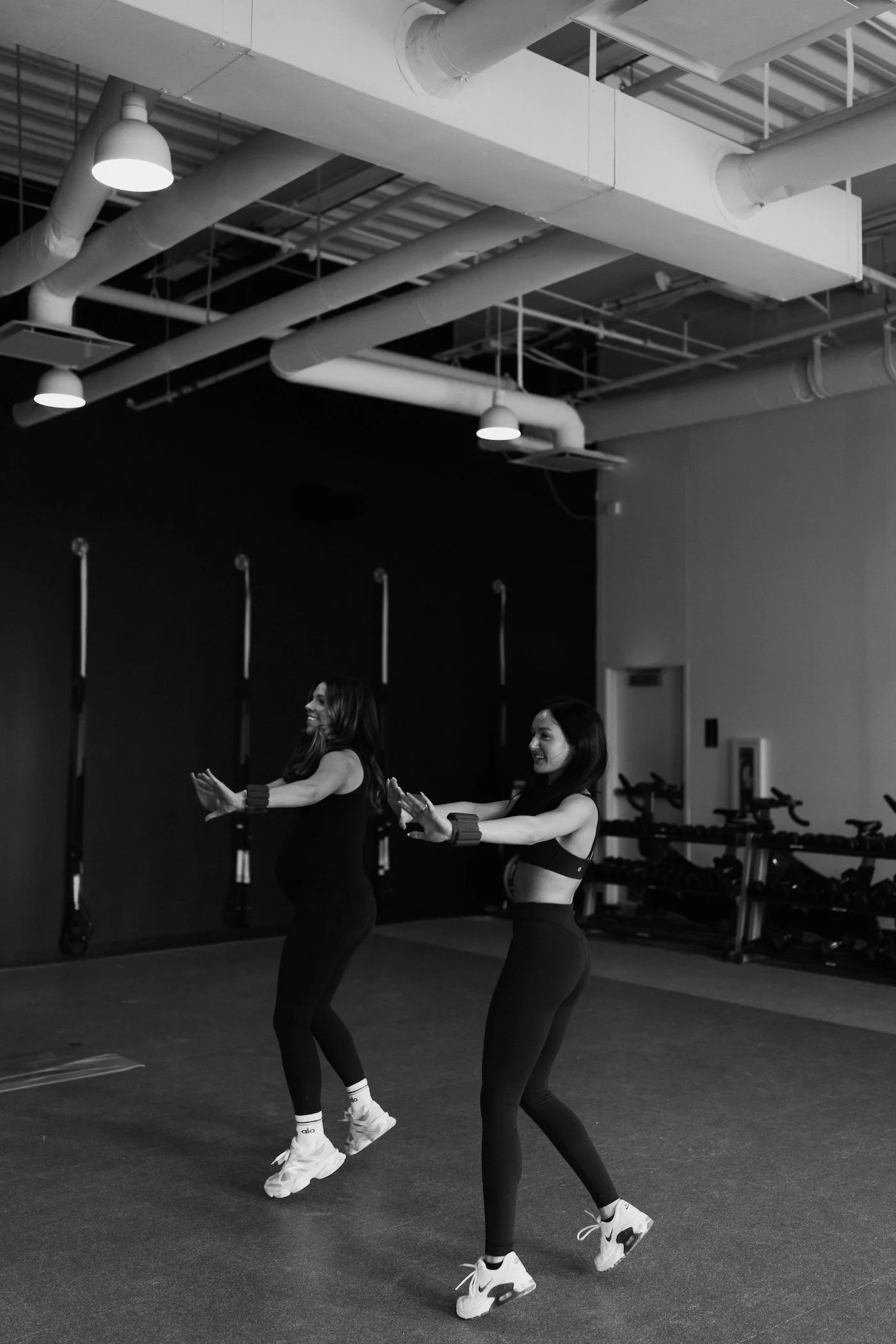 Two women exercising indoors with resistance bands on their wrists, standing on a gym floor in a fitness studio.