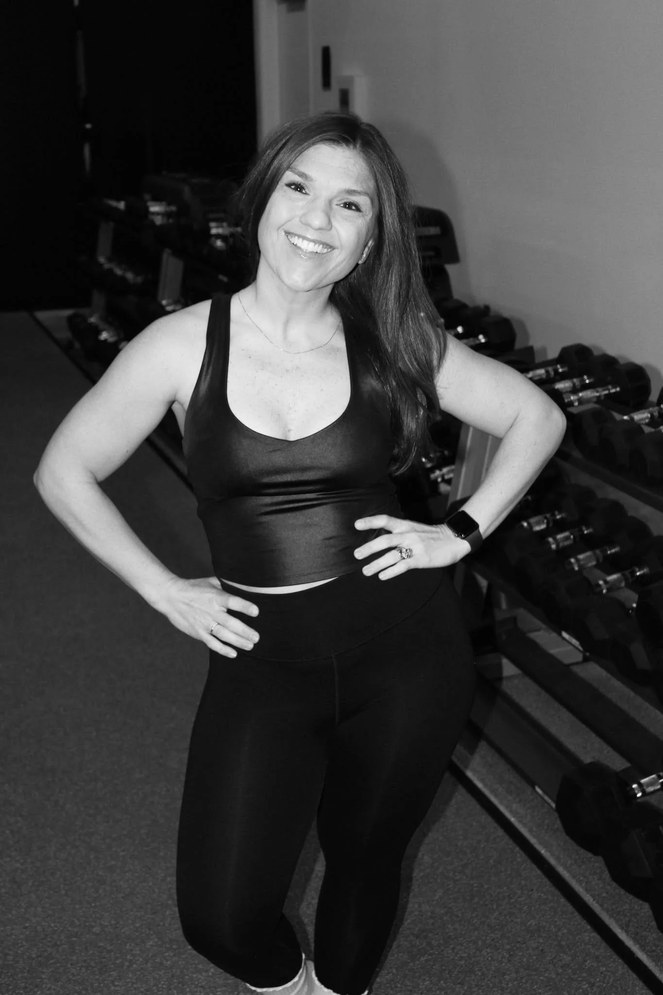 A woman in athletic clothing pointing at the camera and smiling in a gym with dumbbells in the background