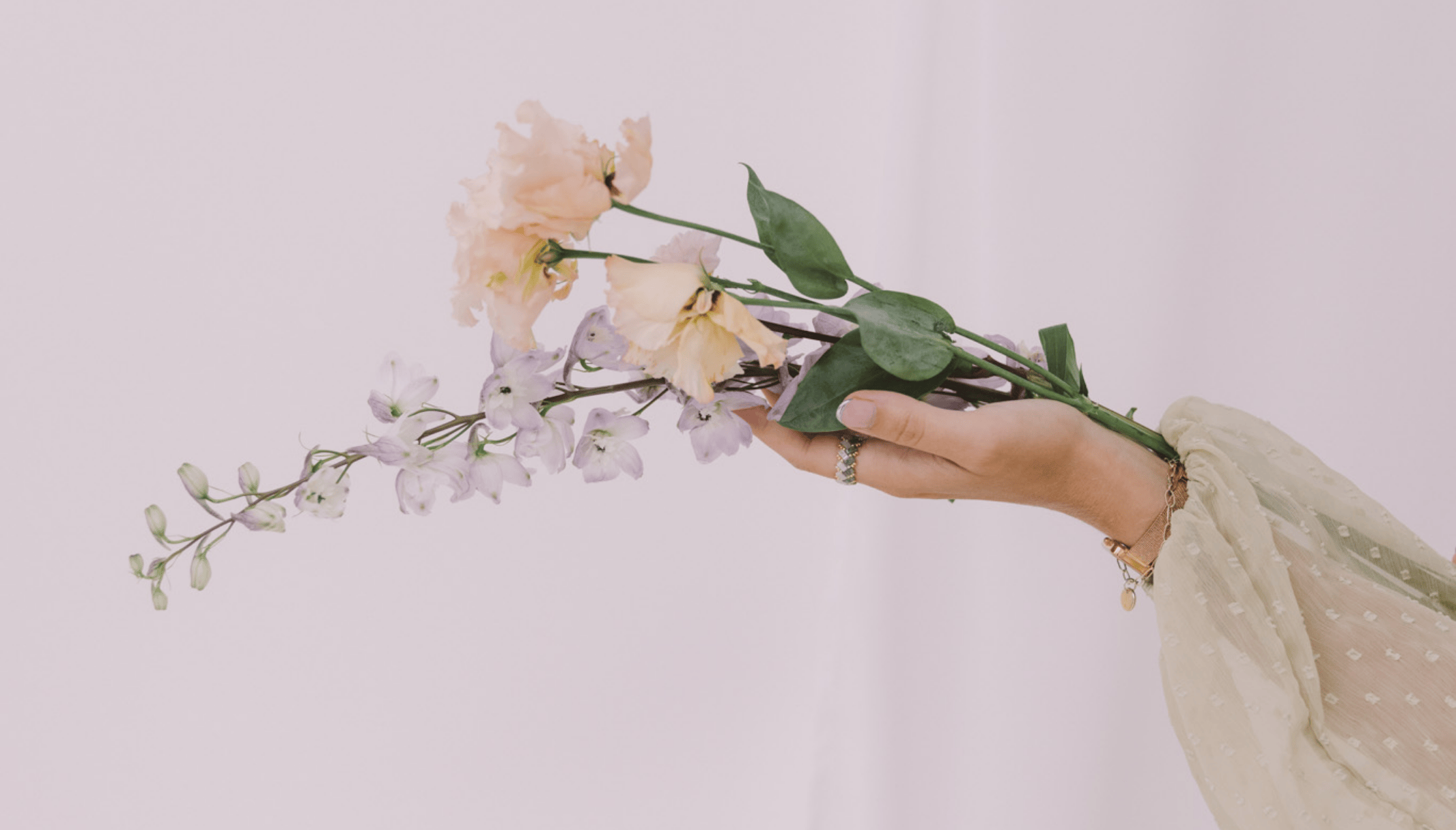Soft, feminine floral arrangement held by hand against a muted neutral background, featuring pastel peach and lilac flowers with an airy, minimal aesthetic.
