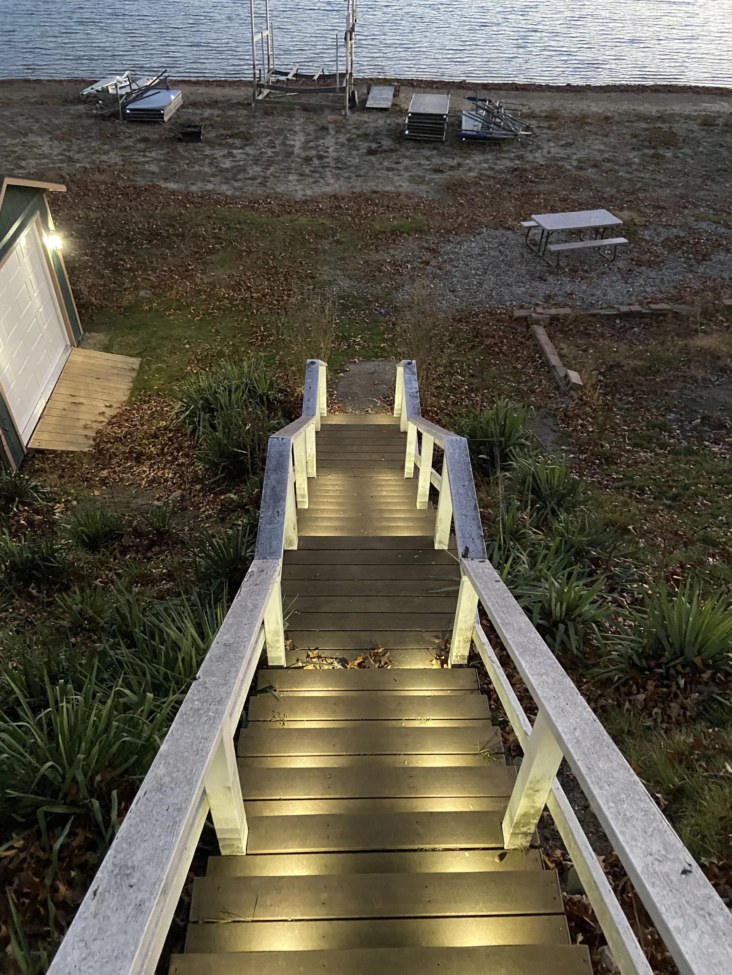 A stepladder staircase with built-in lighting leading down to a sandy beach with a body of water, lounge chairs, and a picnic table in the background.