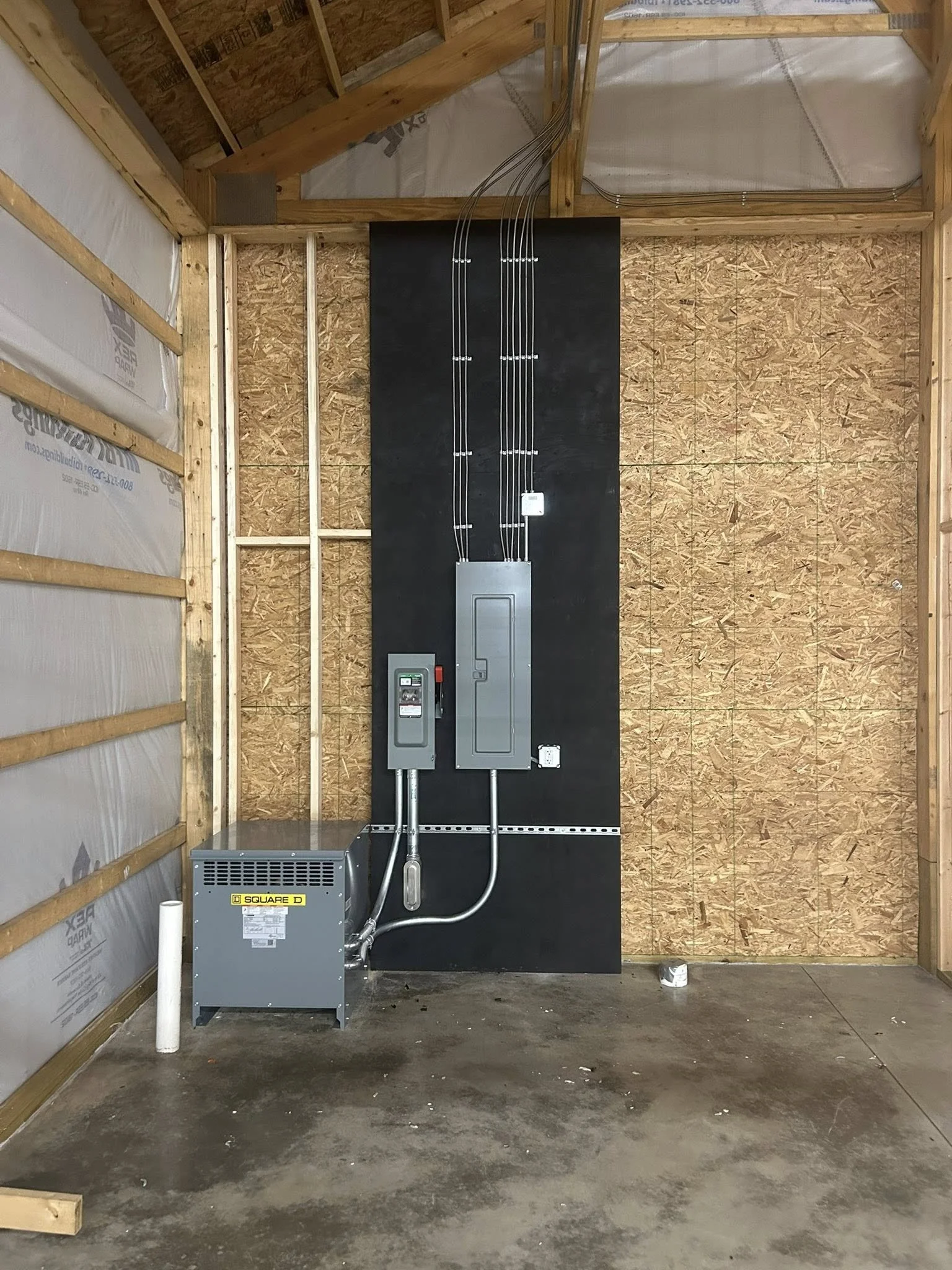 Electrical panel and equipment installed on a black wall inside a partially constructed building with wooden framing and oriented strand board walls.