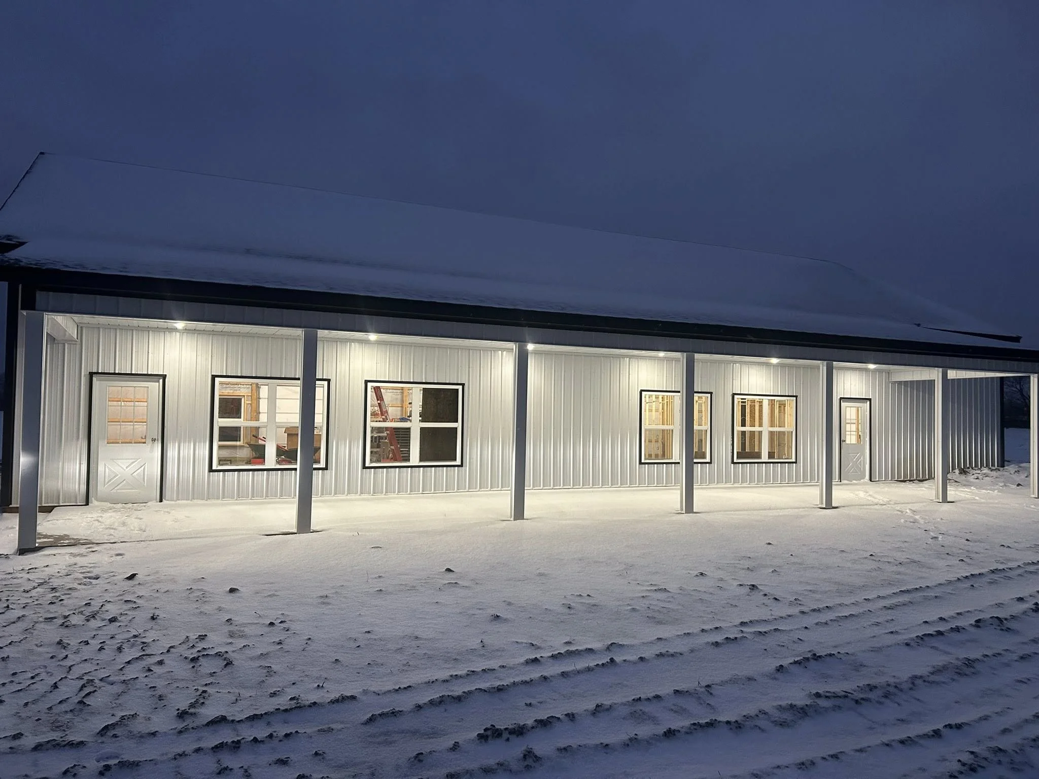 A long, metal-sided building illuminated from inside at night, with snow on the ground and a covered patio area.