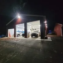 Nighttime view of an automotive workshop with open garage doors, cars inside, and some equipment outside.