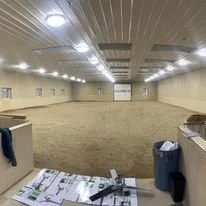 Empty indoor arena with a dirt floor, ceiling lights, beige walls, and some papers and trash in the foreground