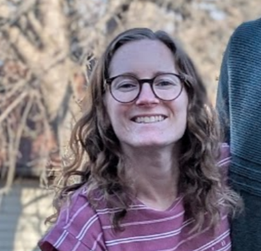 Young woman with curly hair and glasses smiling outdoors.