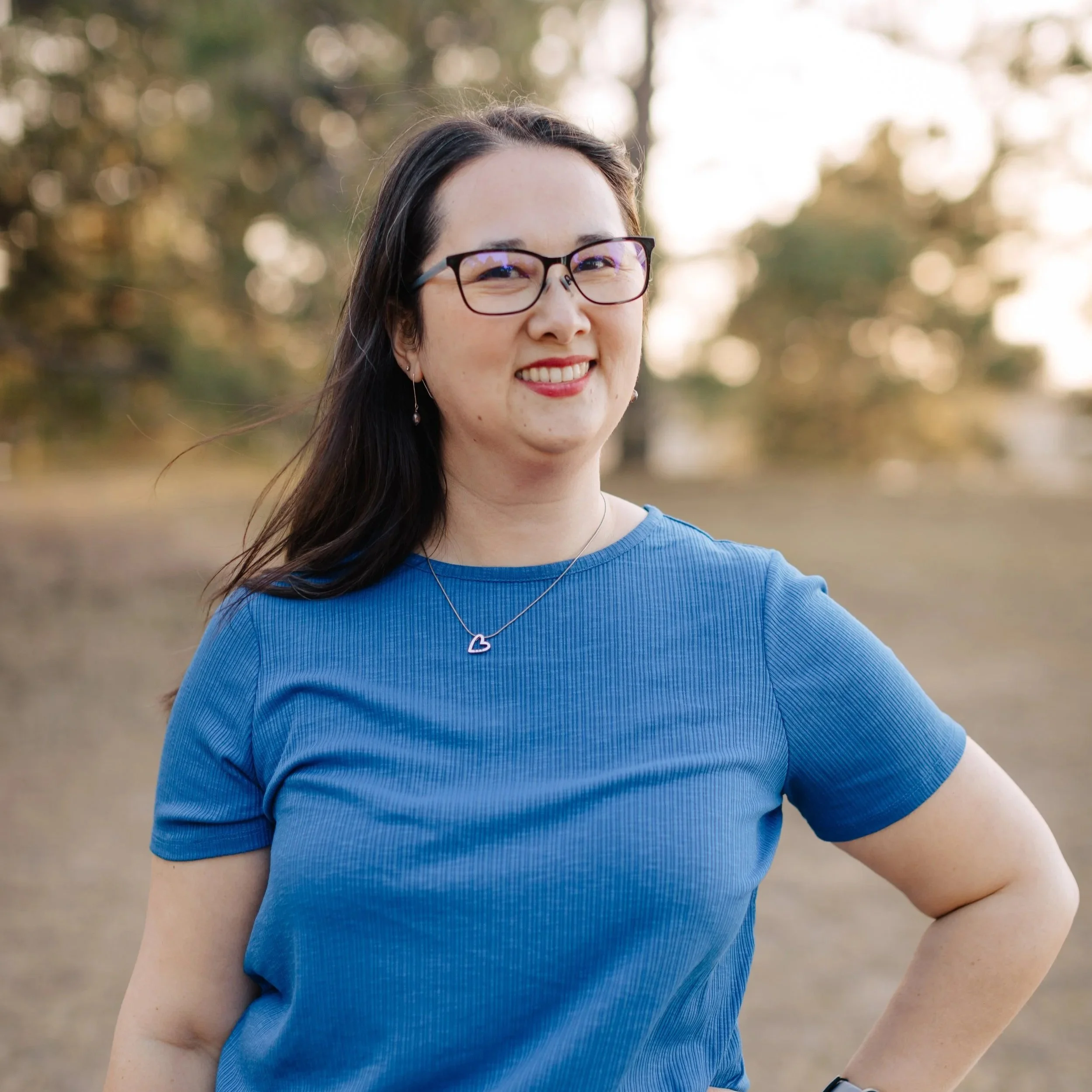 Close-up of a woman smiling outdoors, wearing glasses, a blue shirt, and a heart-shaped necklace, with blurred trees in the background.