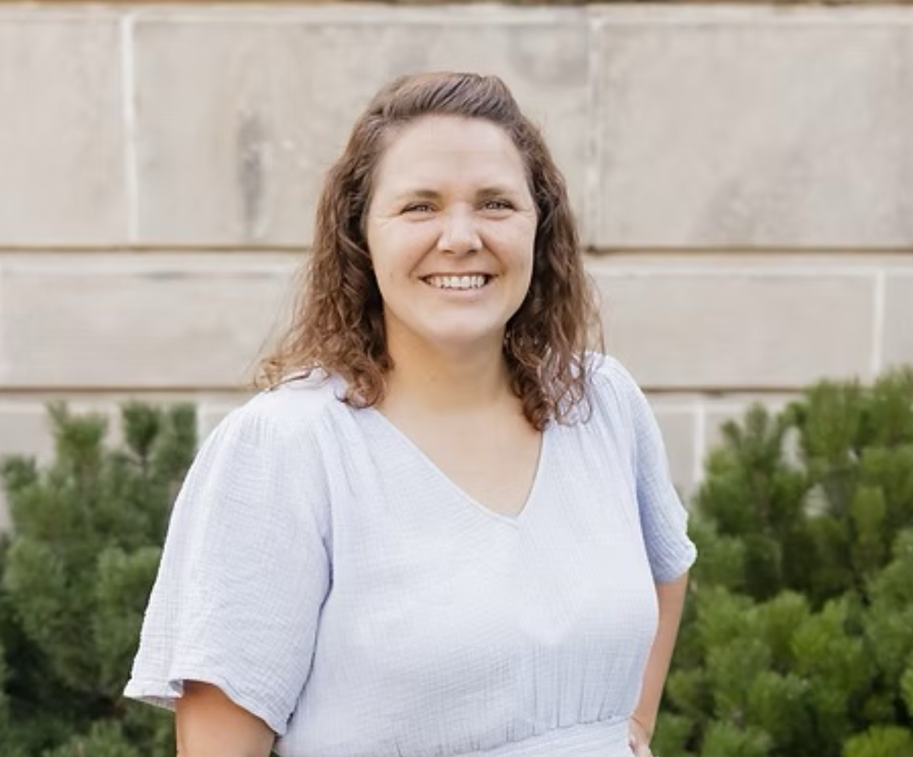 A smiling woman with curly brown hair wearing a light gray top, standing outdoors in front of a stone wall and green bushes.