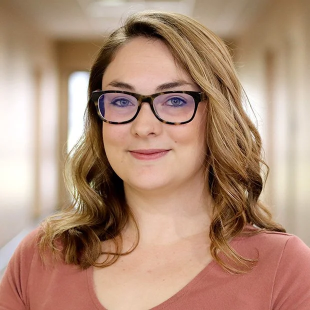 A young woman with wavy brown hair and glasses, smiling, in an indoor setting with blurred background.