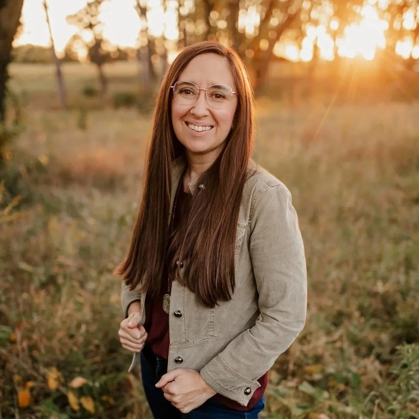 A woman with long brown hair and glasses smiling outdoors during sunset, wearing a beige jacket.