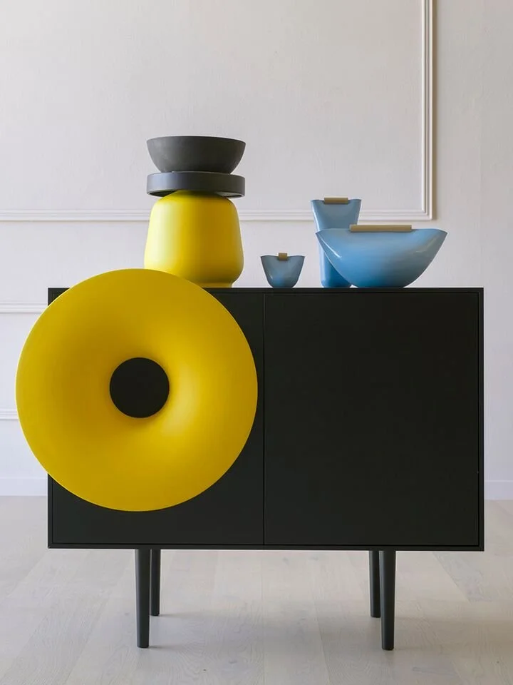 A modern black cabinet with a prominent yellow horn speaker attached to its side, topped with a stack of geometric bowls in gray, yellow, and blue.
