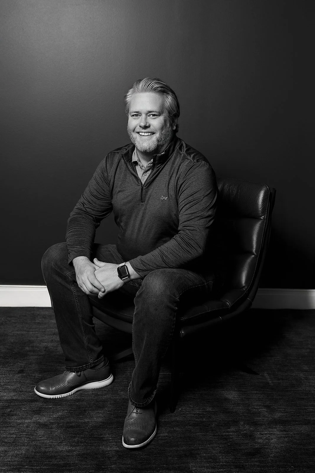 A man with a beard smiling and sitting on an armchair, wearing a blue blazer and a maroon shirt, with abstract artwork on a purple wall in the background.