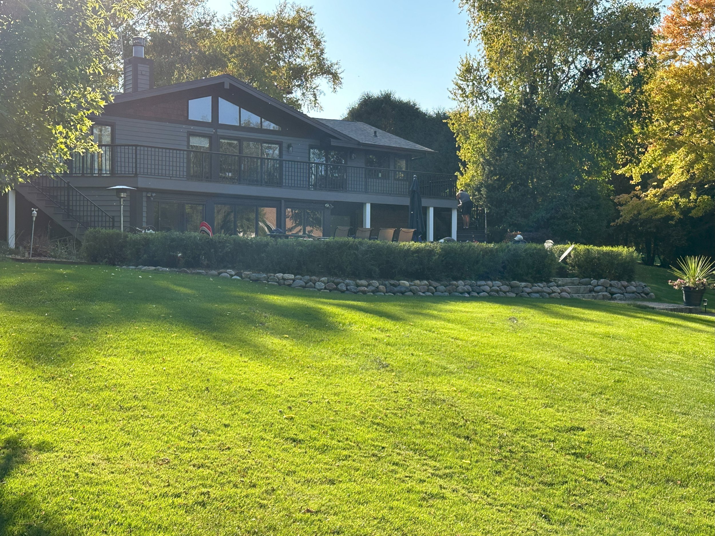 A modern house with large windows and a balcony, surrounded by a well-maintained lawn and trees in a sunny setting.