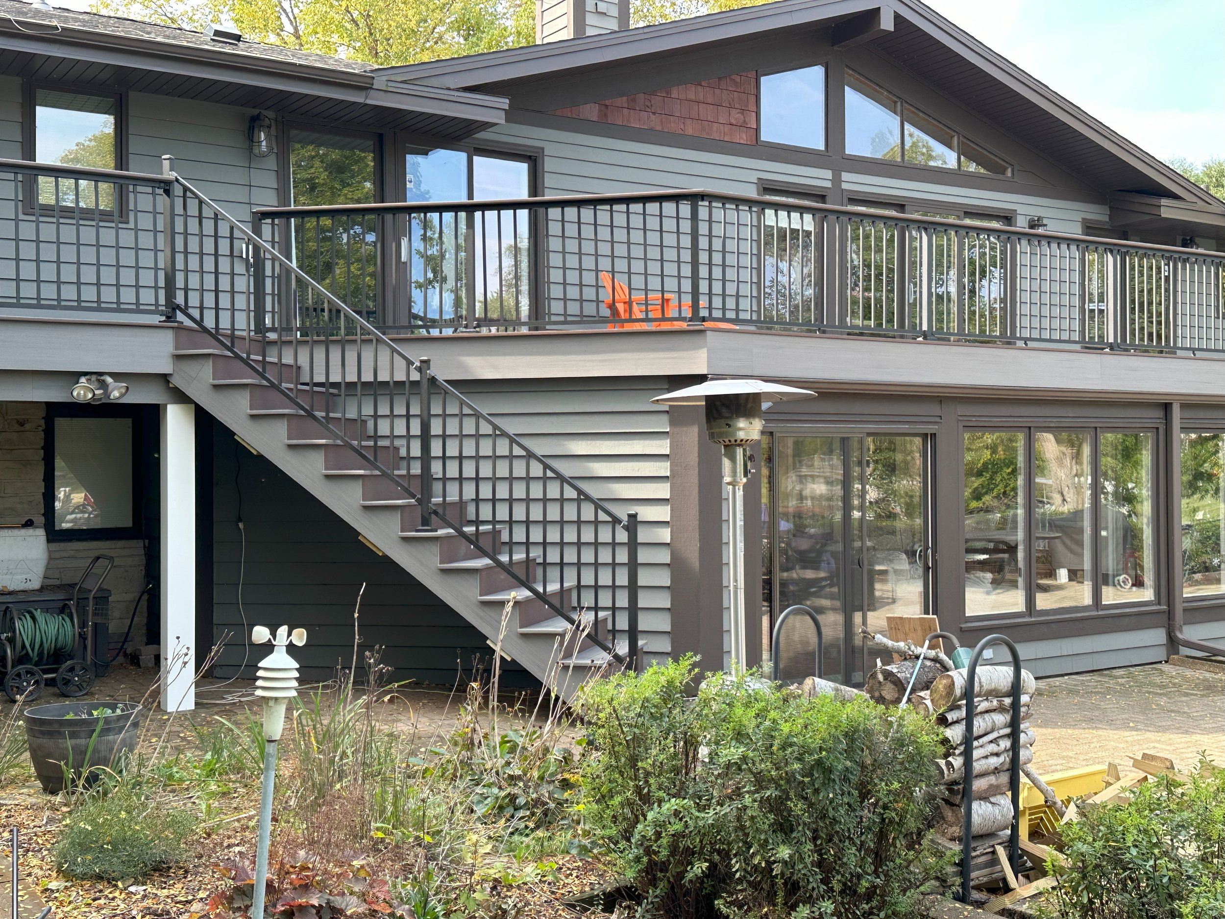 Exterior of a modern house with two stories, featuring large windows and a balcony with a metal railing. There's a staircase leading to the upper level. A patio heater and garden tools are visible. Vegetation and a weather station are in the foregrou