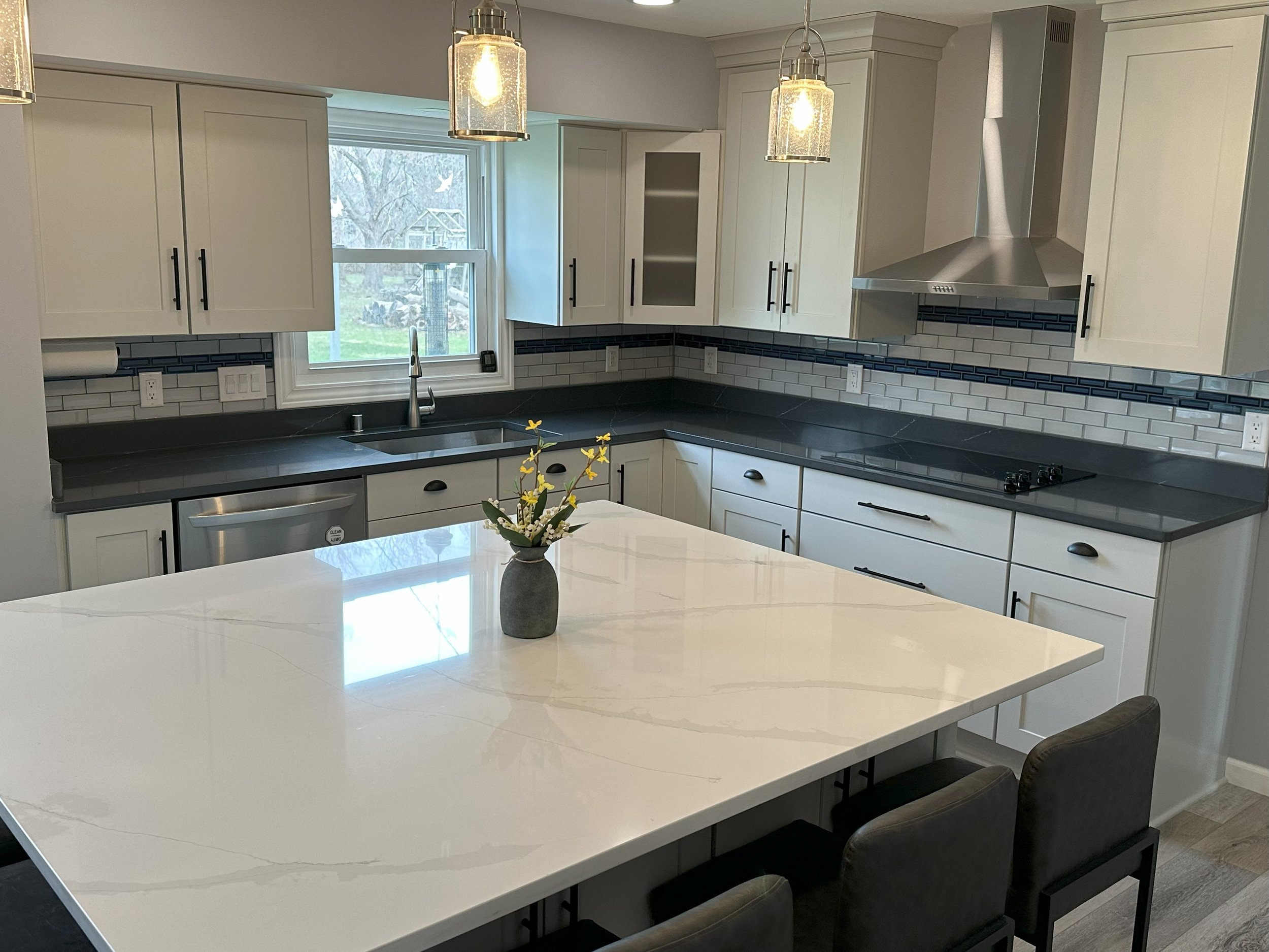 Modern kitchen with white cabinets, gray countertops, stainless steel appliances, large white island with vase, pendant lights, and a tiled backsplash.