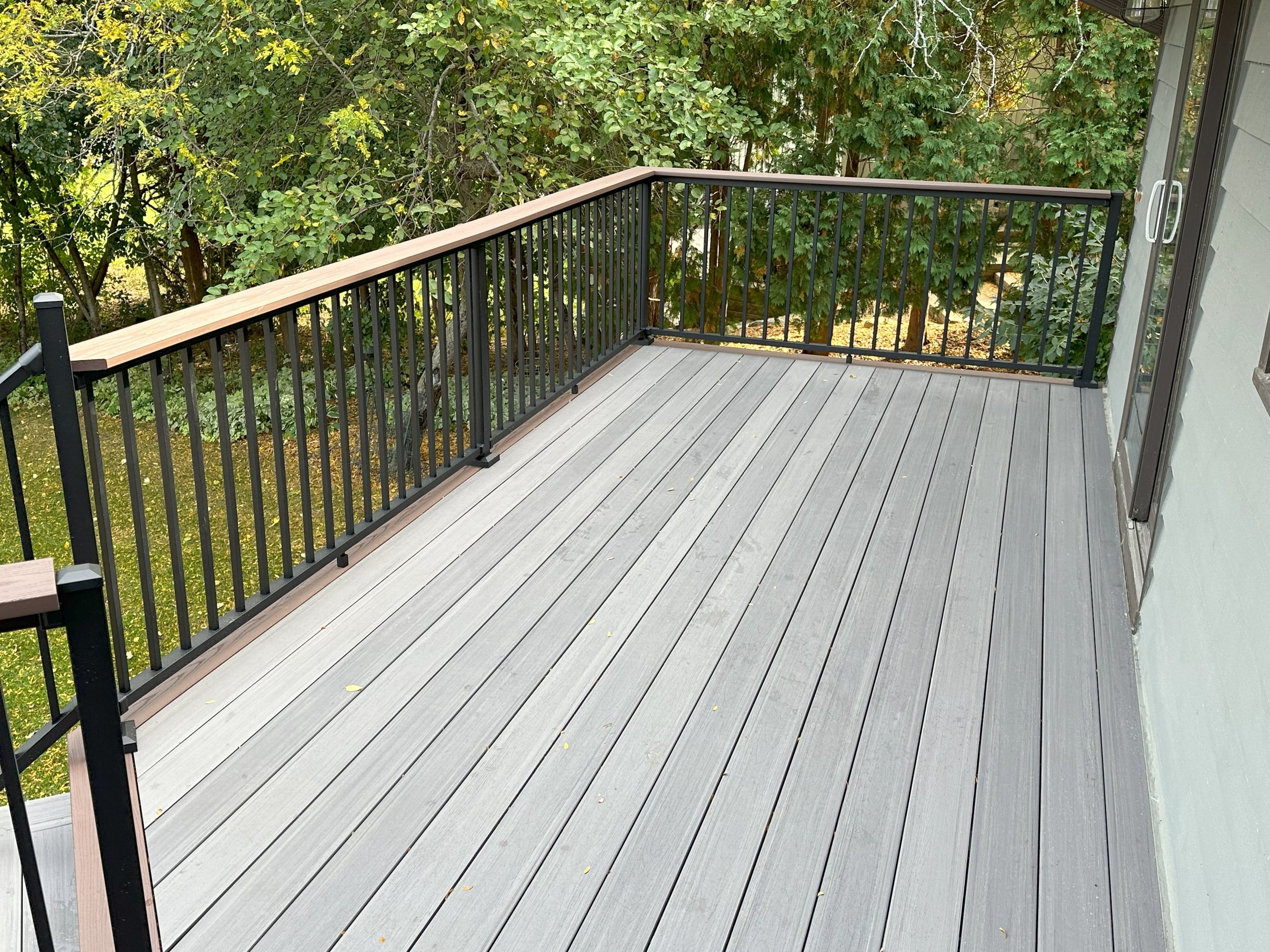 A wooden deck with gray planks and black railings next to a house, surrounded by greenery.