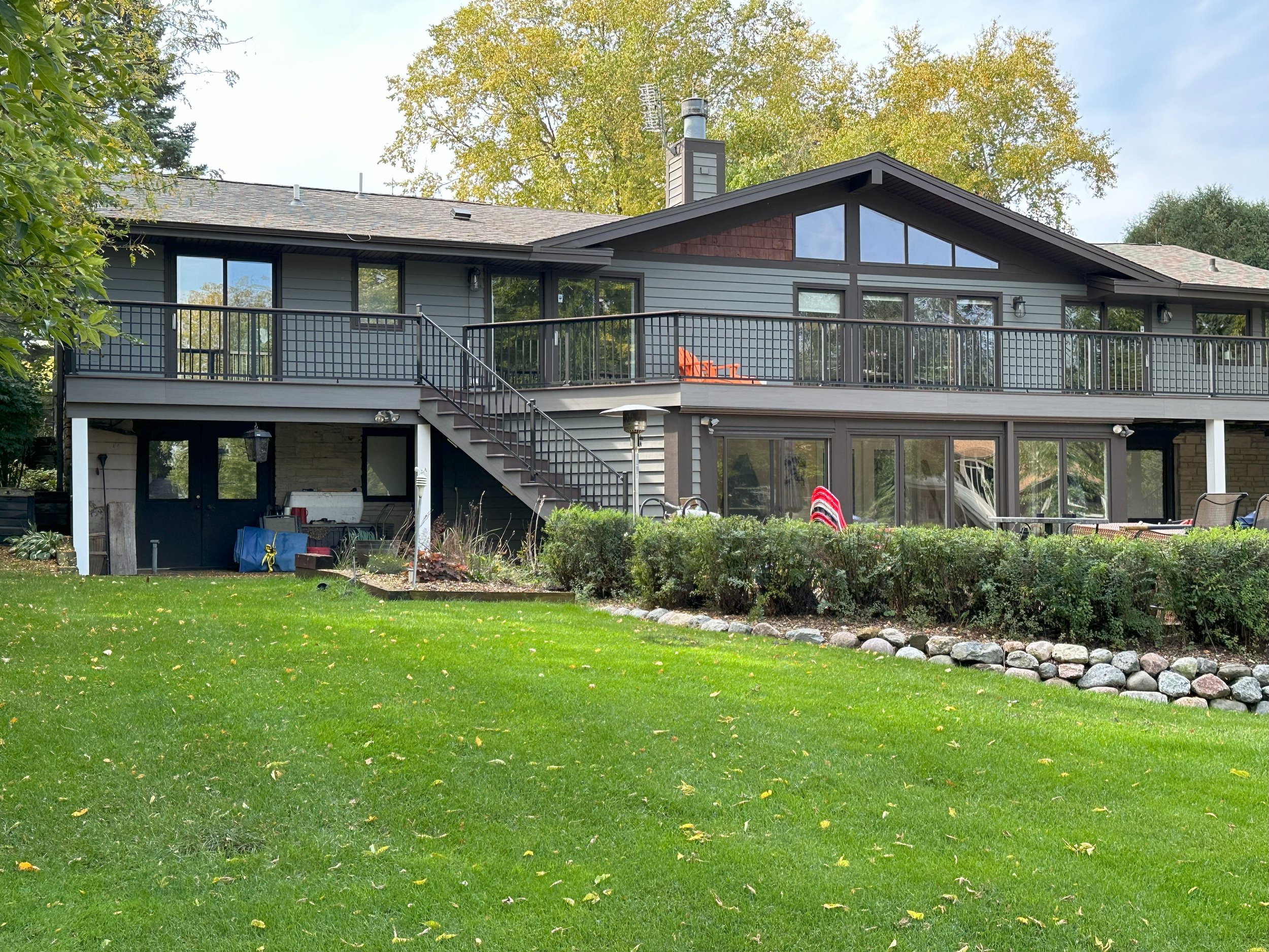 Modern two-story house with large windows and a wraparound balcony, surrounded by green lawn and trees, with a stone-lined garden in the foreground.
