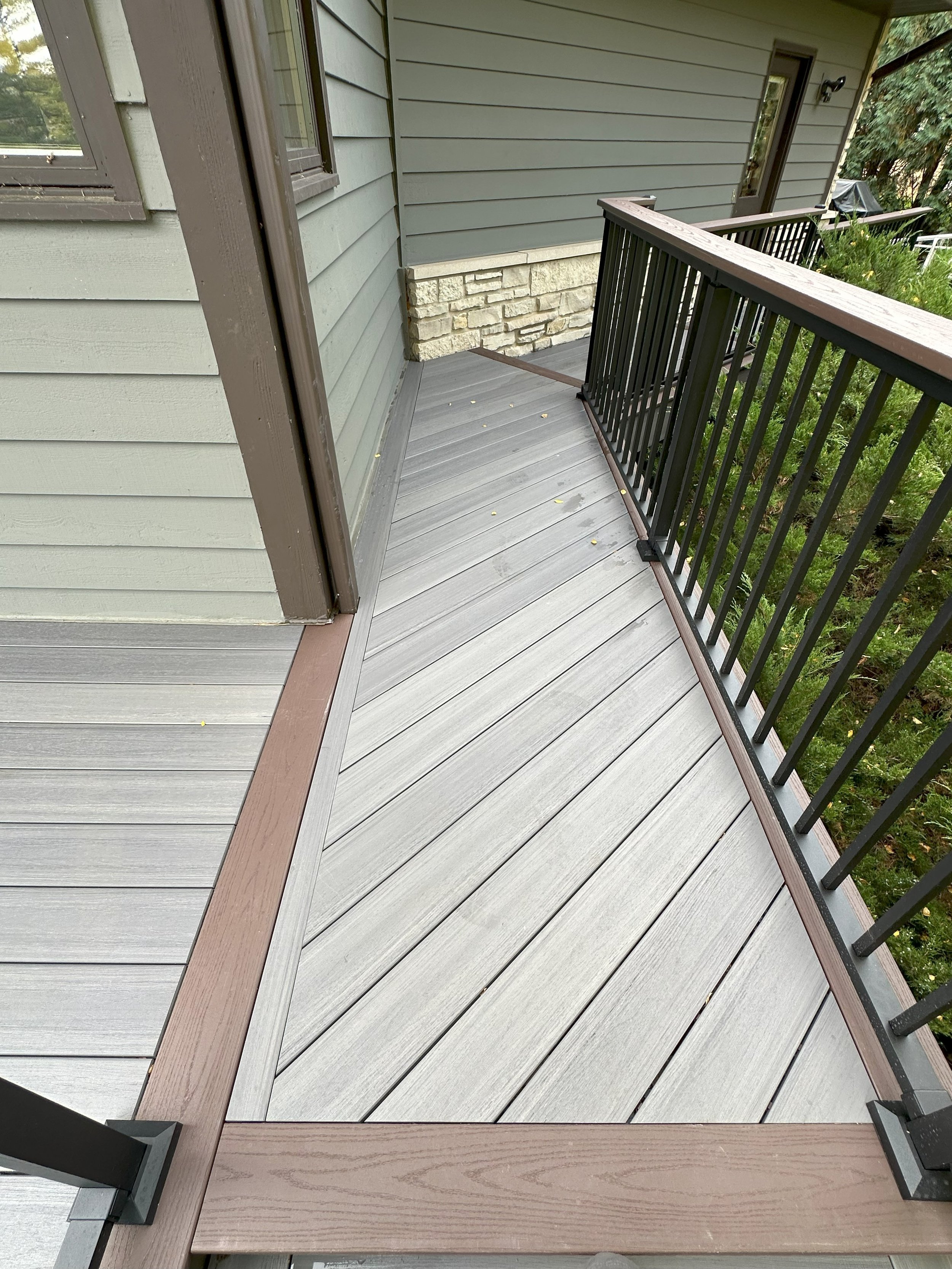 Narrow wooden deck pathway with gray composite planks, brown borders, and a metal railing alongside a house with blue-green siding and stone accents.