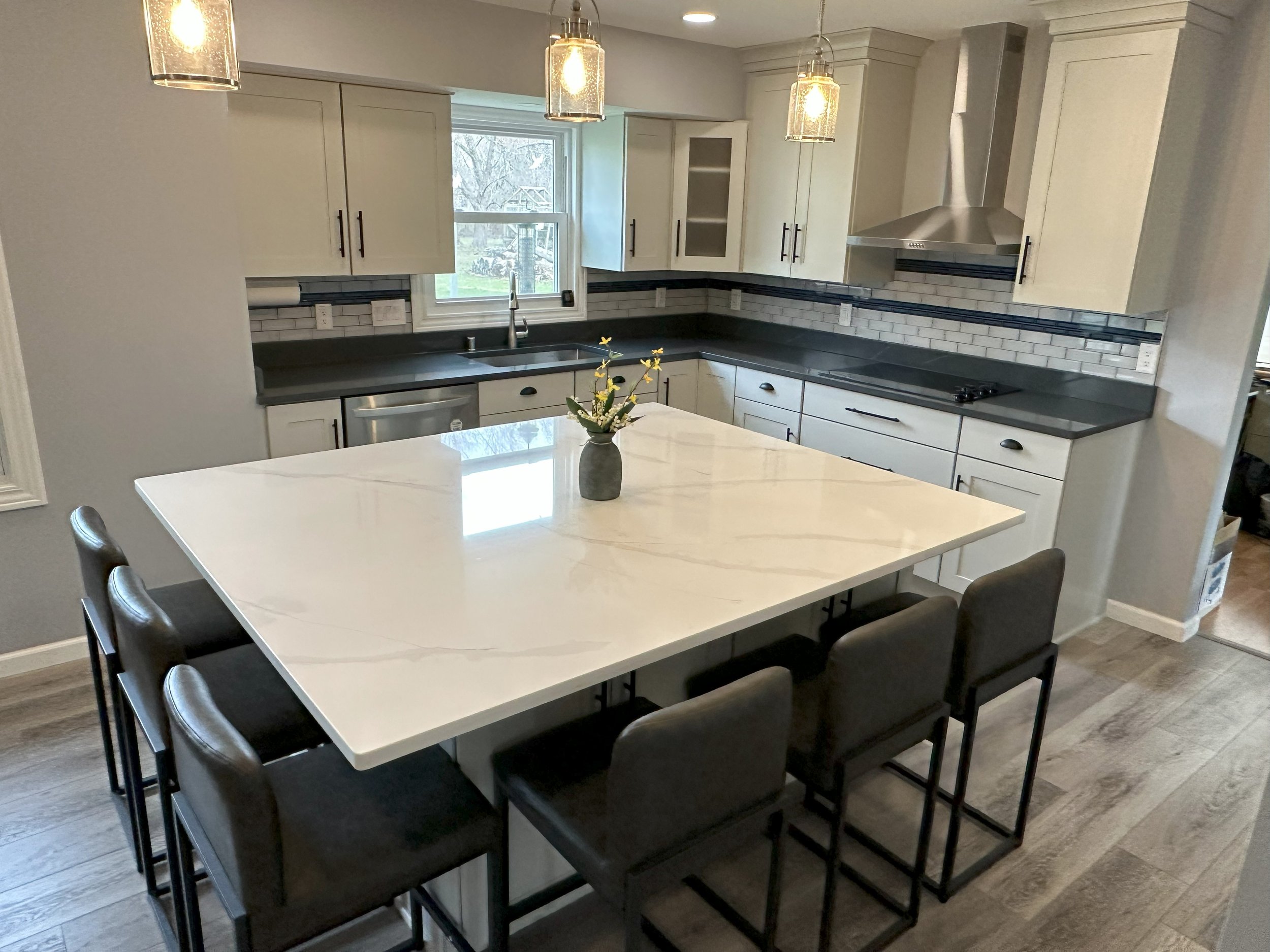 Modern kitchen with a large white marble island, black counter stools, stainless steel appliances, subway tile backsplash, pendant lights, gray cabinets, and wood flooring.