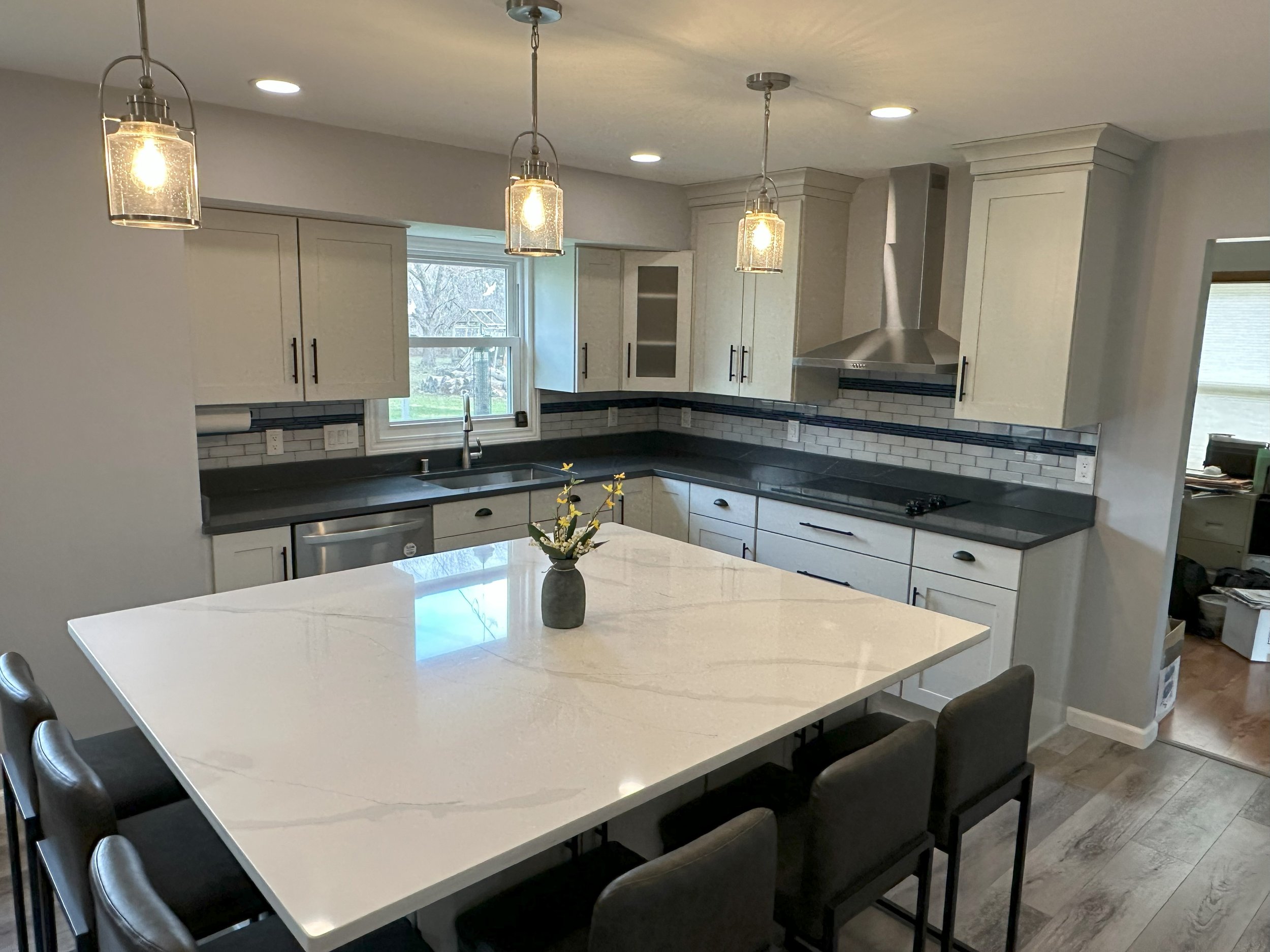 Modern kitchen with white cabinets, dark countertops, and a large marble island. Pendant lights hang above the island with stools. Stainless steel appliances are present, including a range hood and dishwasher. The backsplash features white subway til