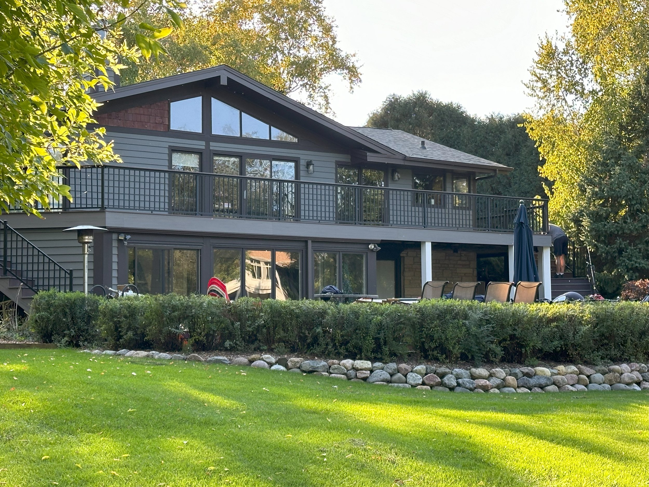 Modern two-story house with dark wood panels and large windows, surrounded by greenery and a manicured lawn. Outdoor seating area and stone-bordered hedges are in the foreground.