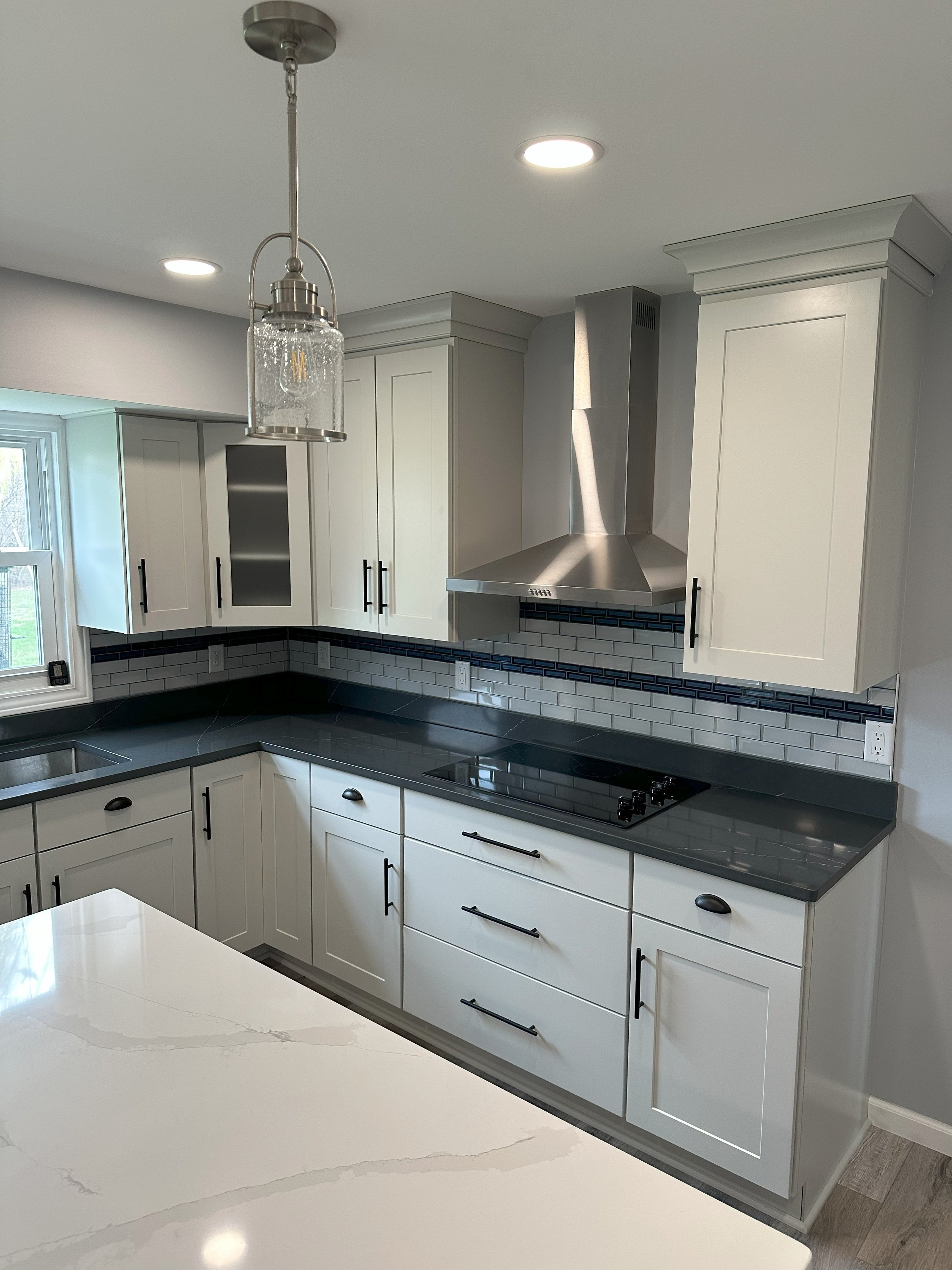 Modern kitchen with white cabinets, dark countertops, a stainless steel hood, and pendant lighting. Features a tiled backsplash and an island with a marble-like surface.