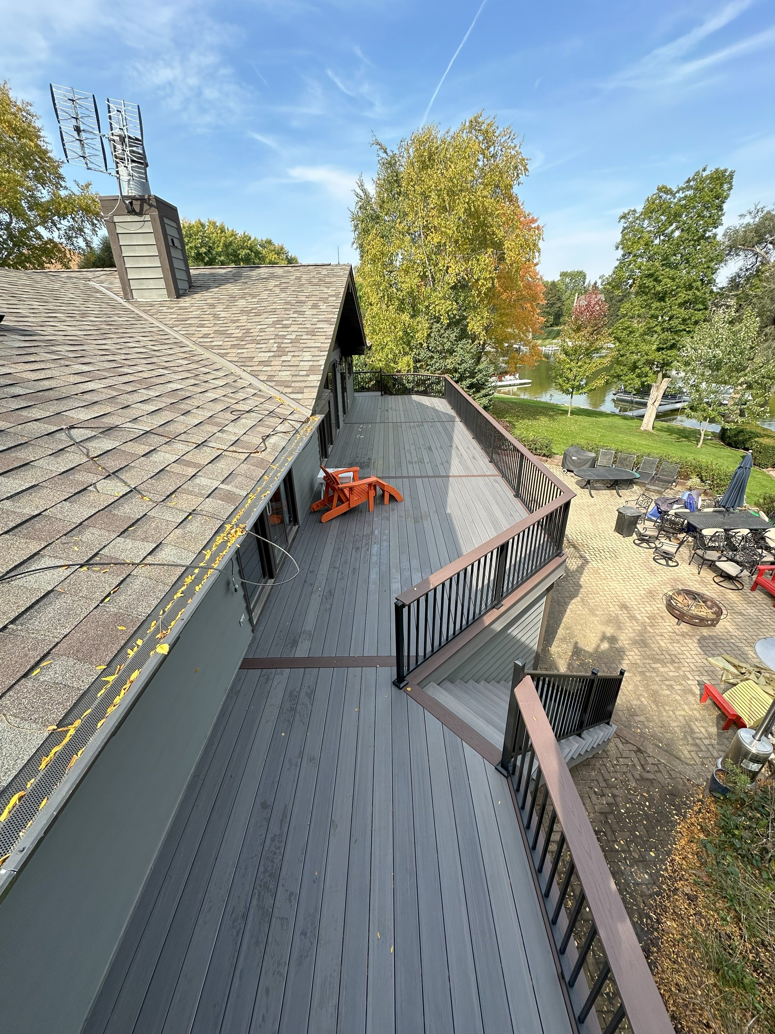 Elevated view of a rooftop deck attached to a house with chairs and railing, overlooking a backyard with trees and a water body in the background on a sunny day.