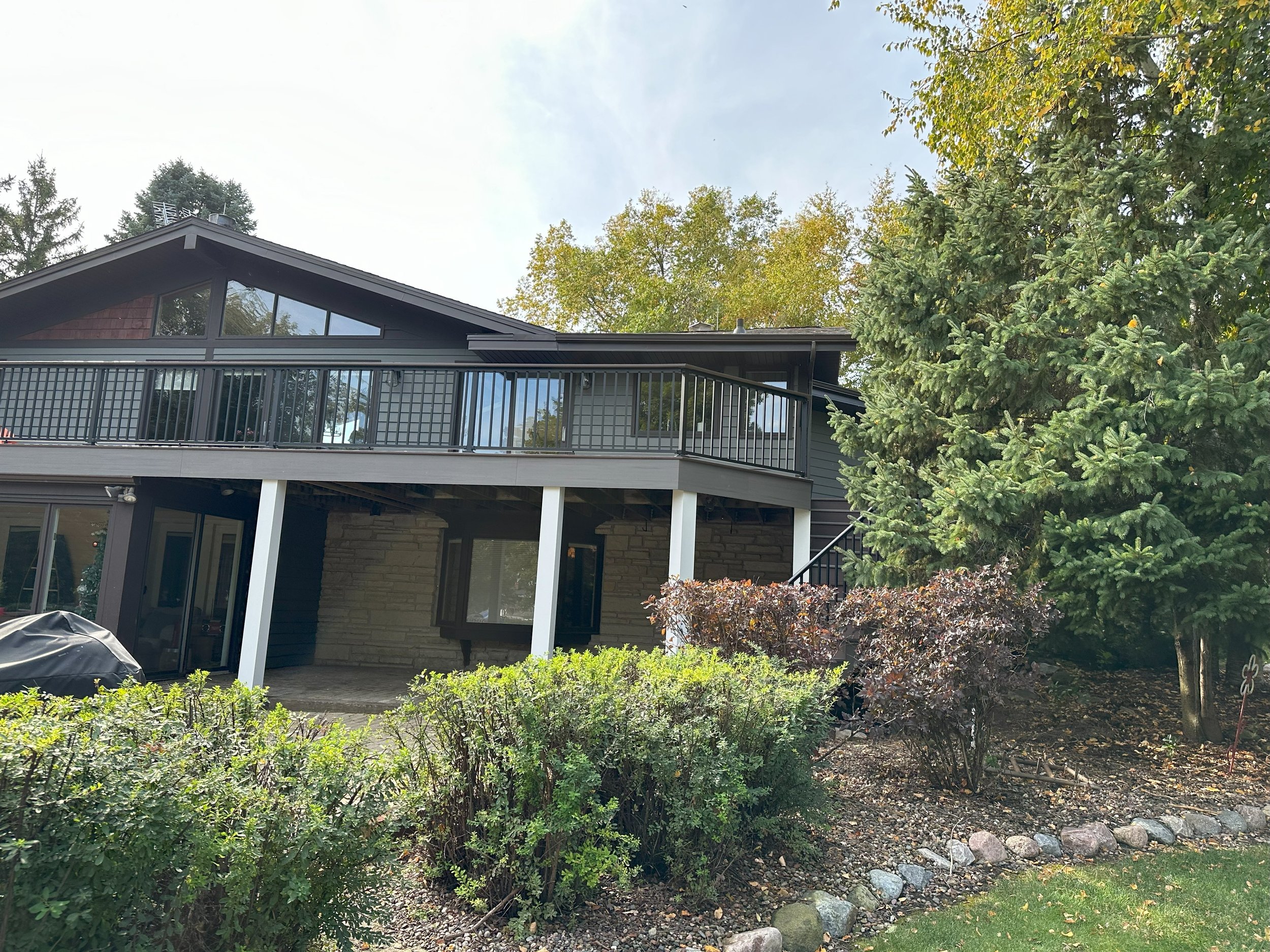 A modern house with a large deck and railing, surrounded by trees and shrubs in a landscaped yard.