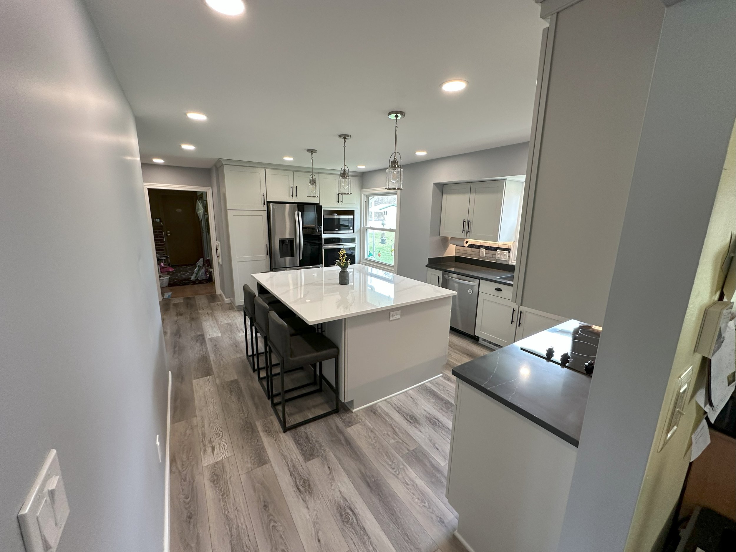 Modern kitchen with white cabinets, stainless steel appliances, and a large island with bar stools. The room features recessed lighting, light gray walls, and wood vinyl flooring.