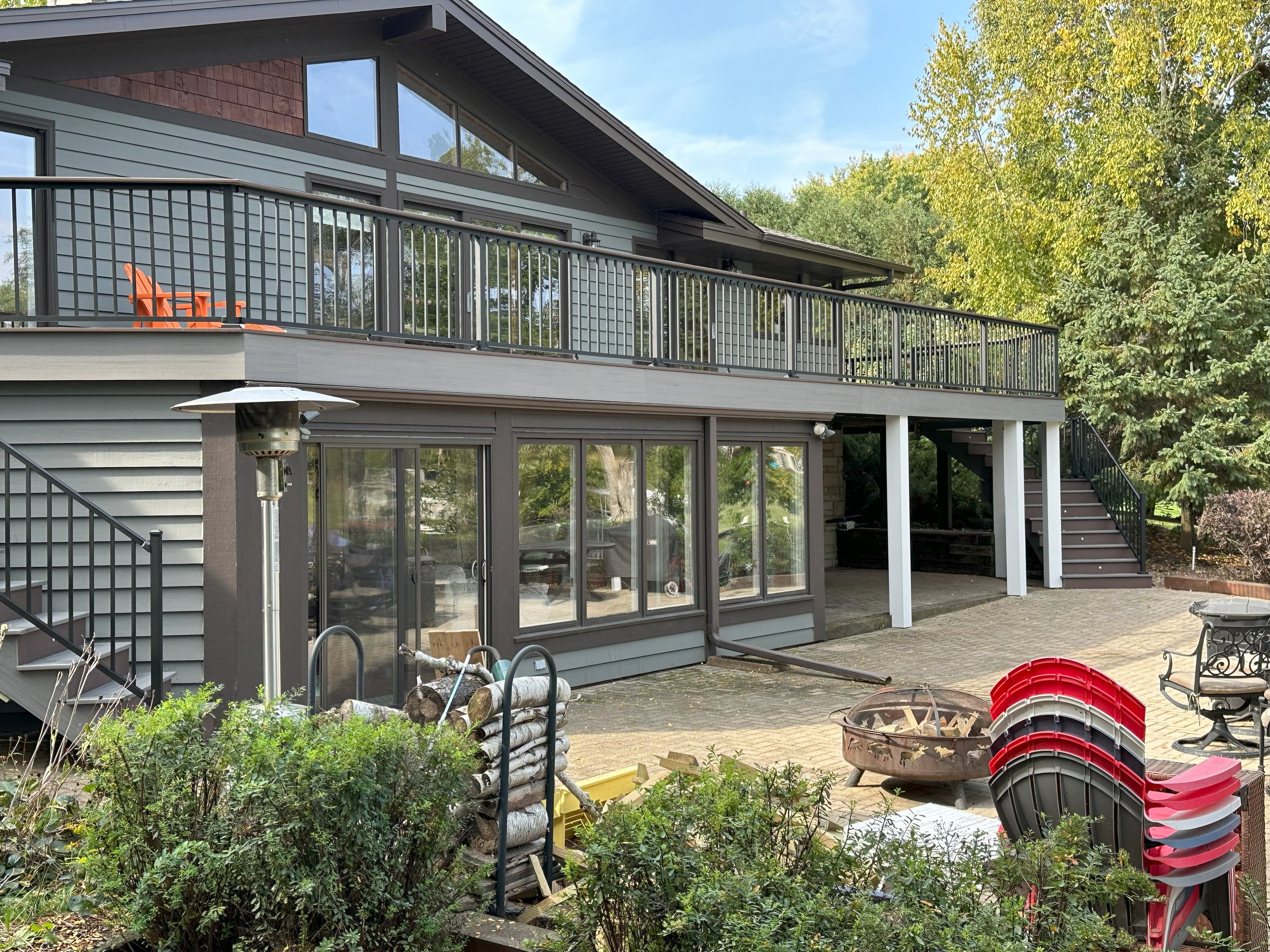Modern two-story house with large windows, spacious balcony, outdoor seating, and stacked firewood on a brick patio surrounded by trees.