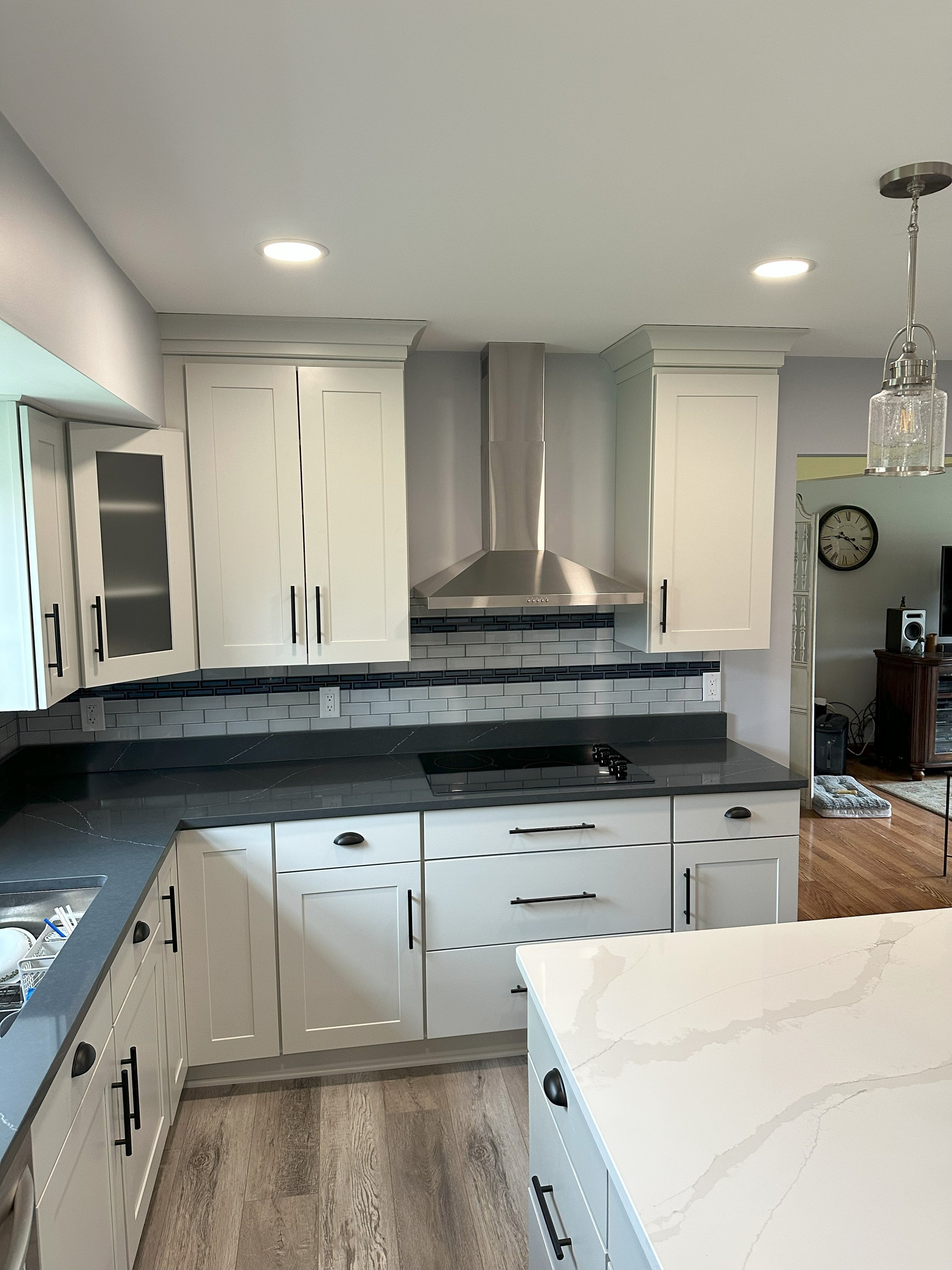 Modern kitchen with white cabinetry, stainless steel range hood, dark countertops, and gray subway tile backsplash. Includes an island with marble-like surface and pendant lighting.