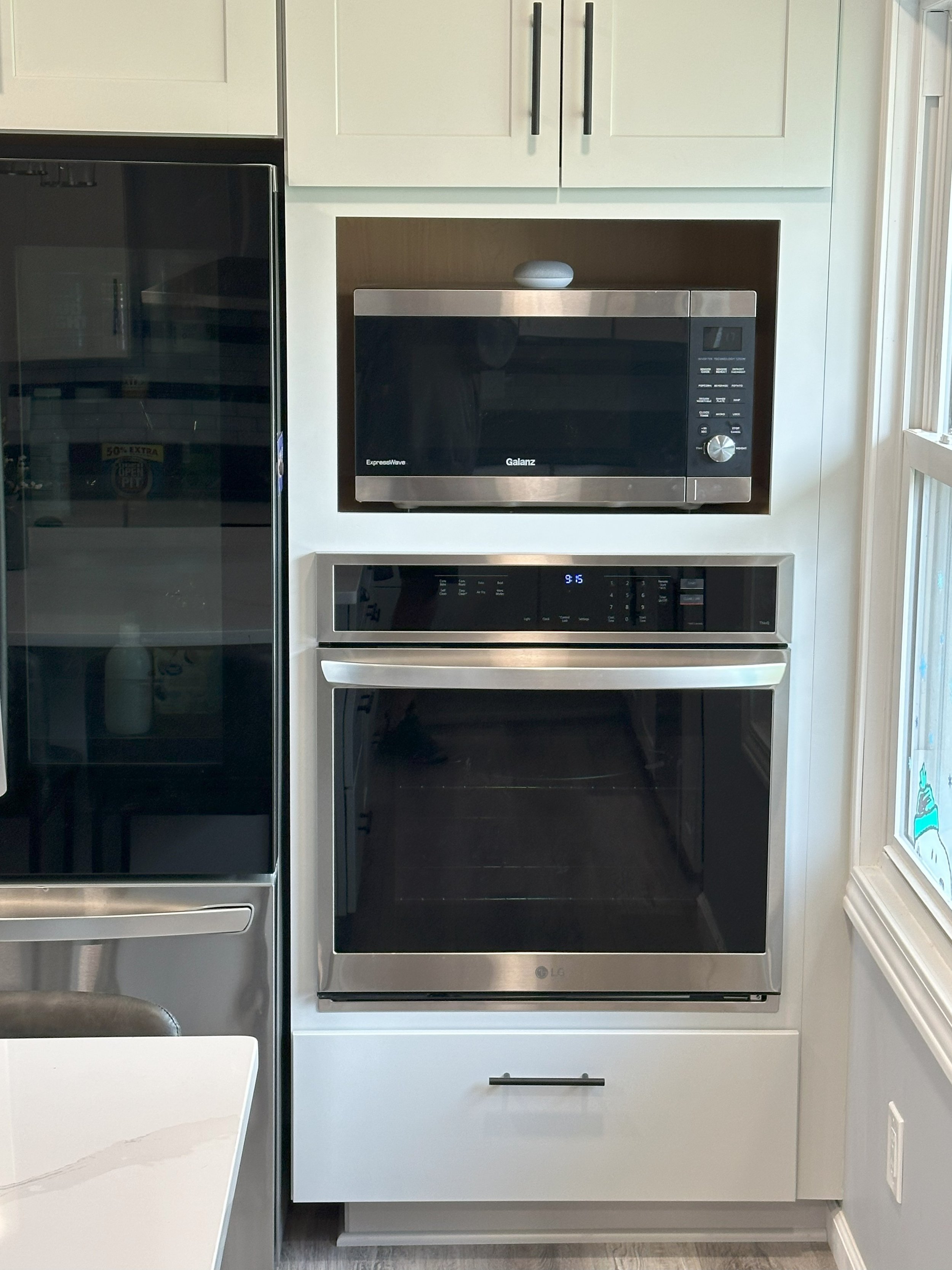 Built-in kitchen appliances featuring a stainless steel microwave above an oven, with white cabinetry and a nearby refrigerator.