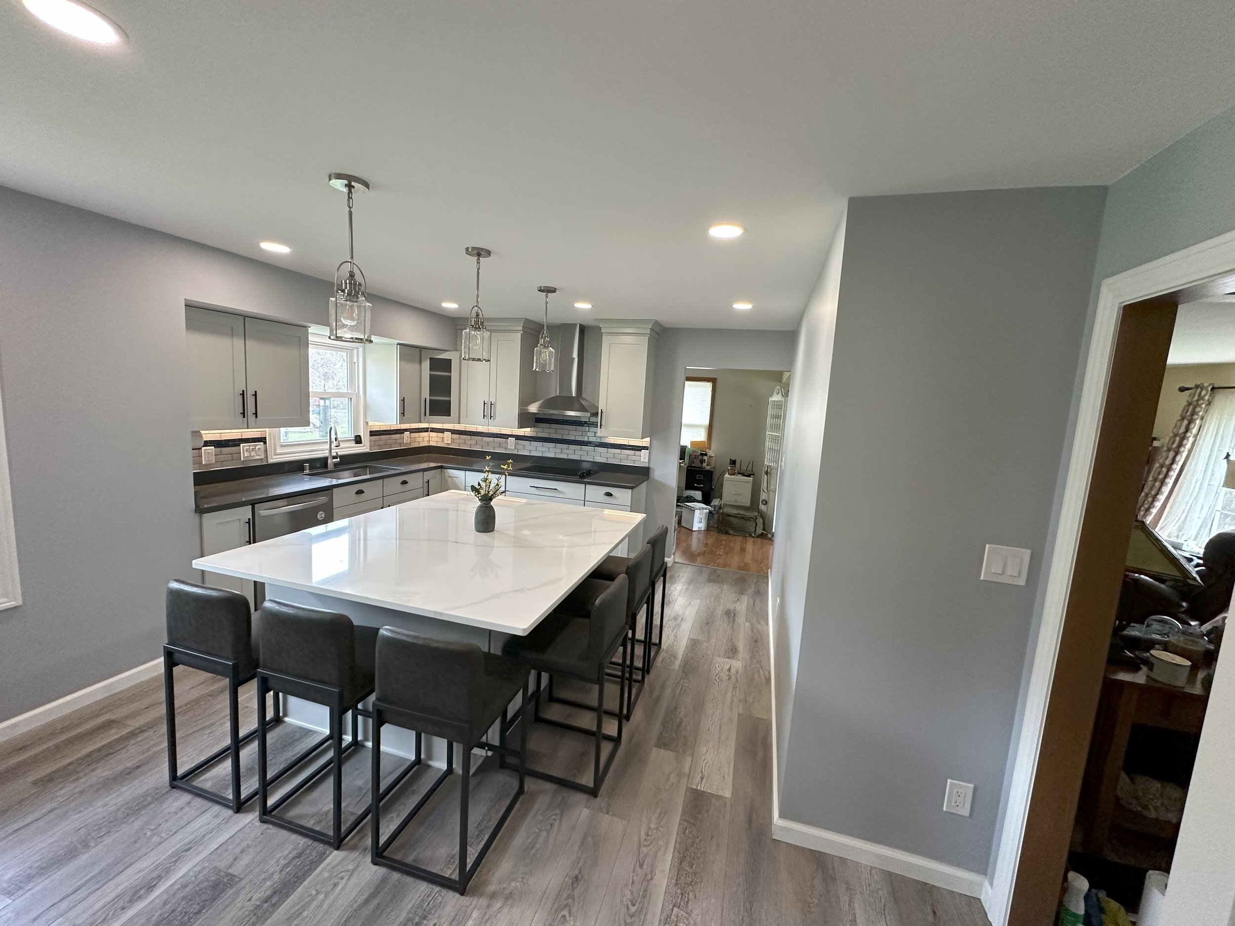 Modern kitchen with large white island, dark chairs, pendant lights, gray cabinets, and wood flooring.
