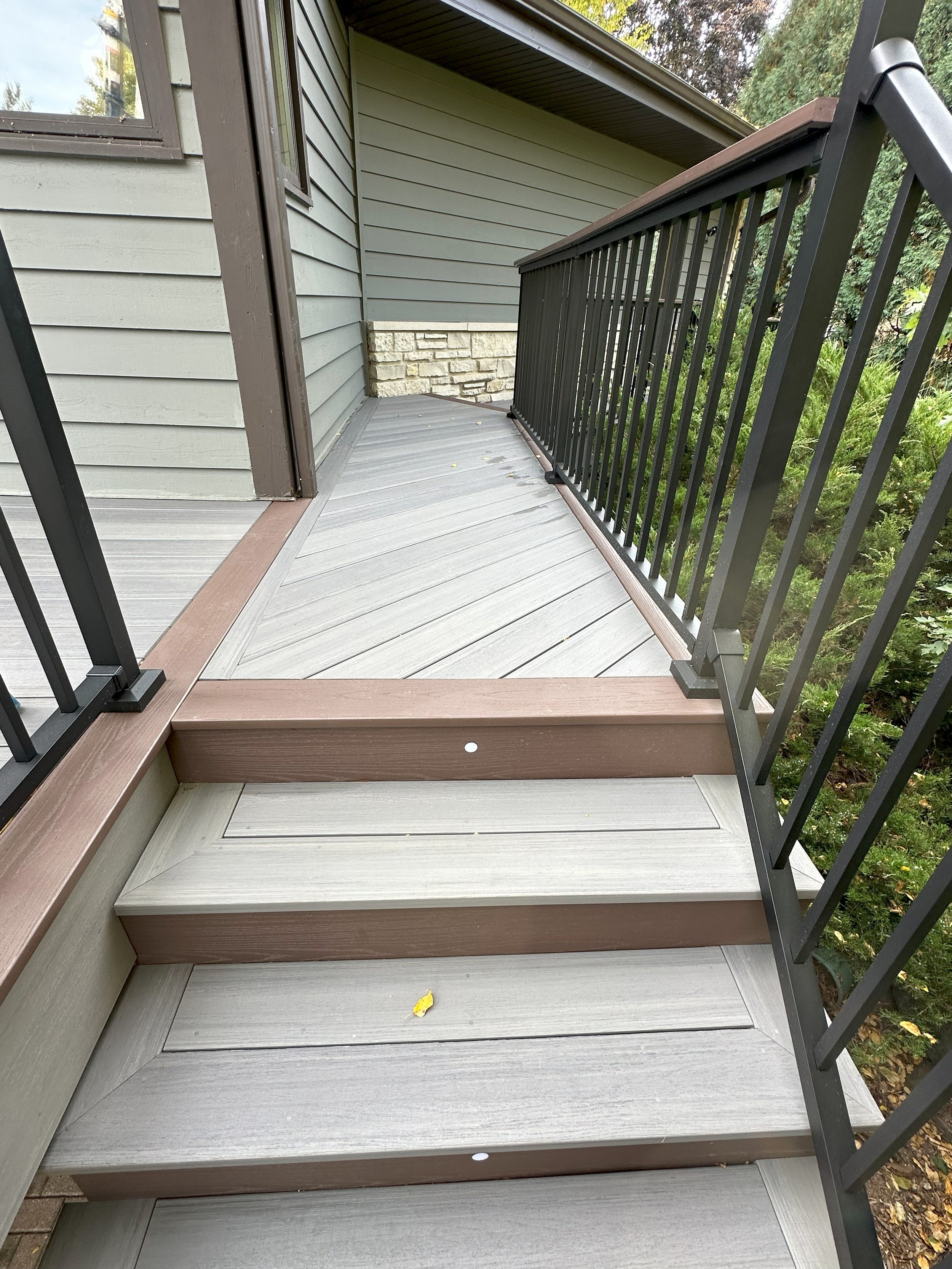 Outdoor deck with stairs and railing, adjacent to a house wall with siding and brickwork, surrounded by greenery.