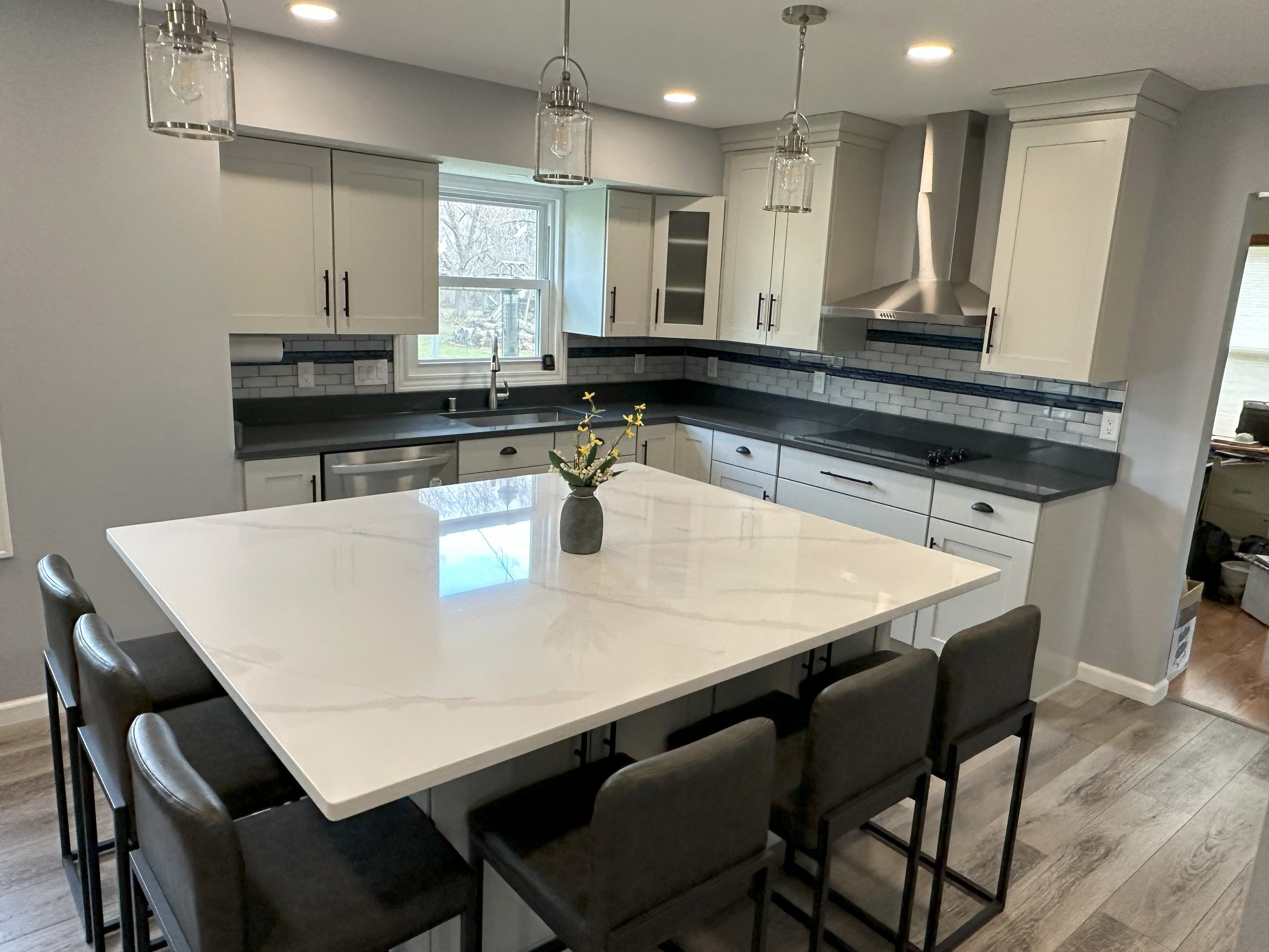 Modern kitchen with white cabinets, large central island with marble countertop, bar stools, stainless steel appliances, and pendant lighting. A vase with flowers is on the island.