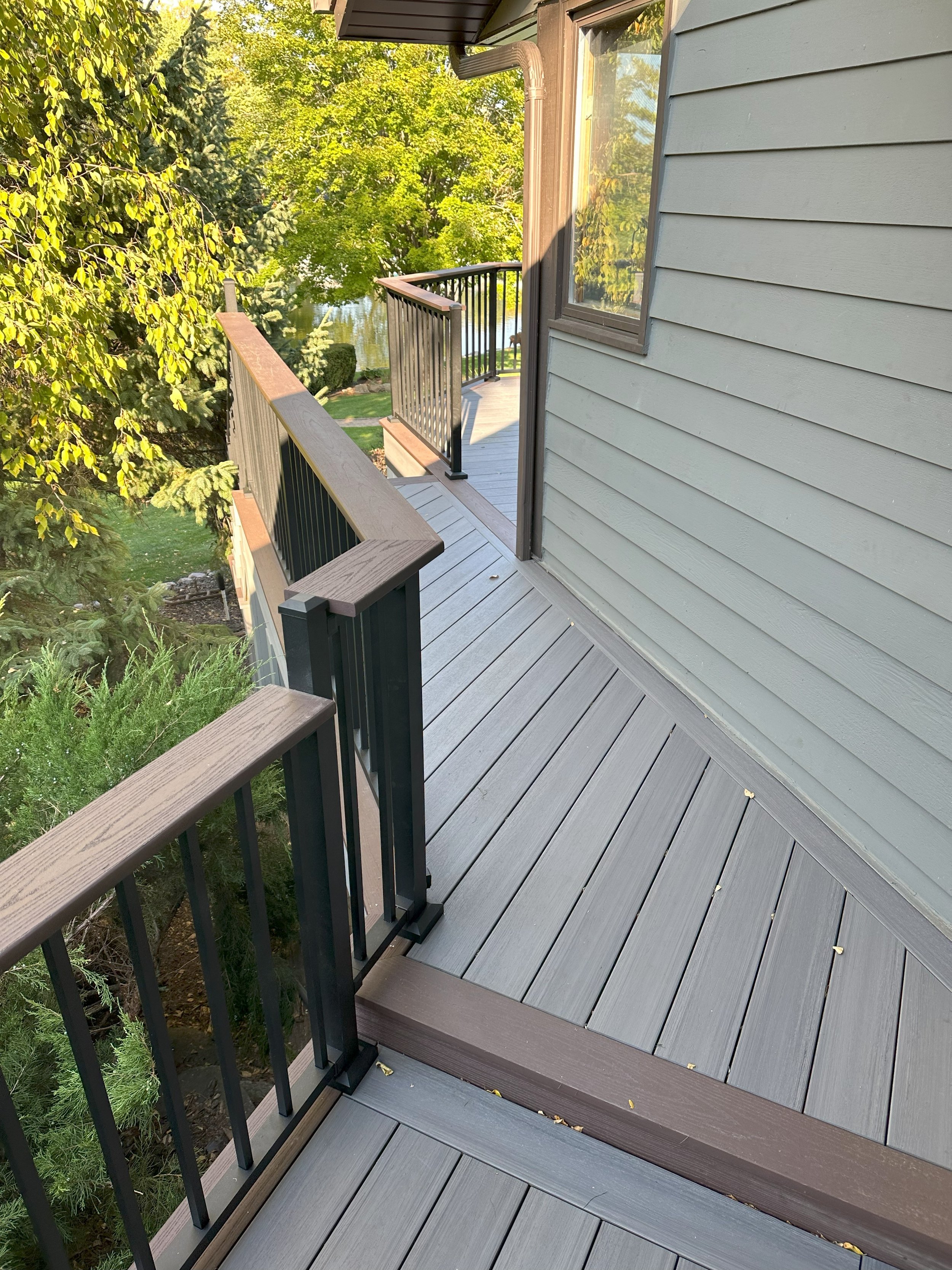 A wooden deck with railings connected to a house, surrounded by trees and greenery.