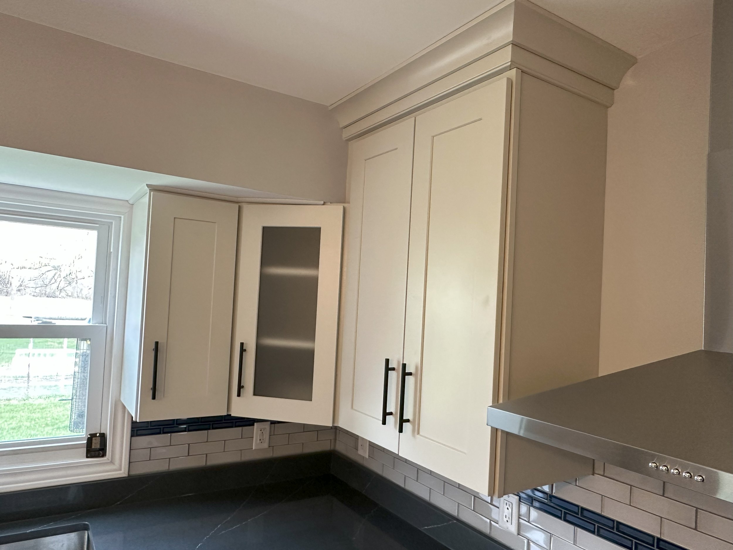 Modern kitchen corner with beige cabinets, black handles, under-cabinet lighting, and tiled backsplash.