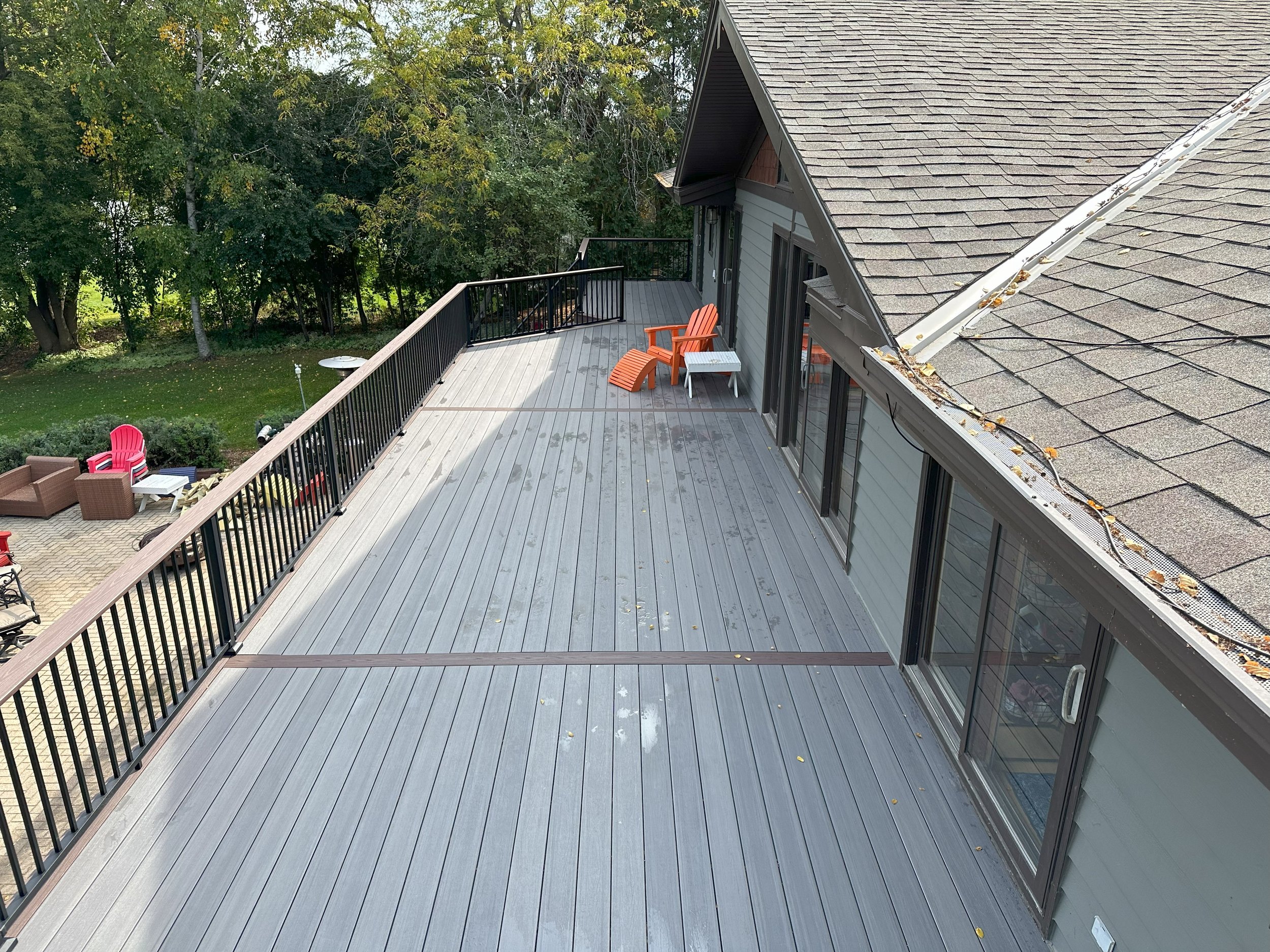Large gray wooden deck attached to a house, with black railings and two orange chairs. Trees and a patio area with red chairs and a table are visible in the background.