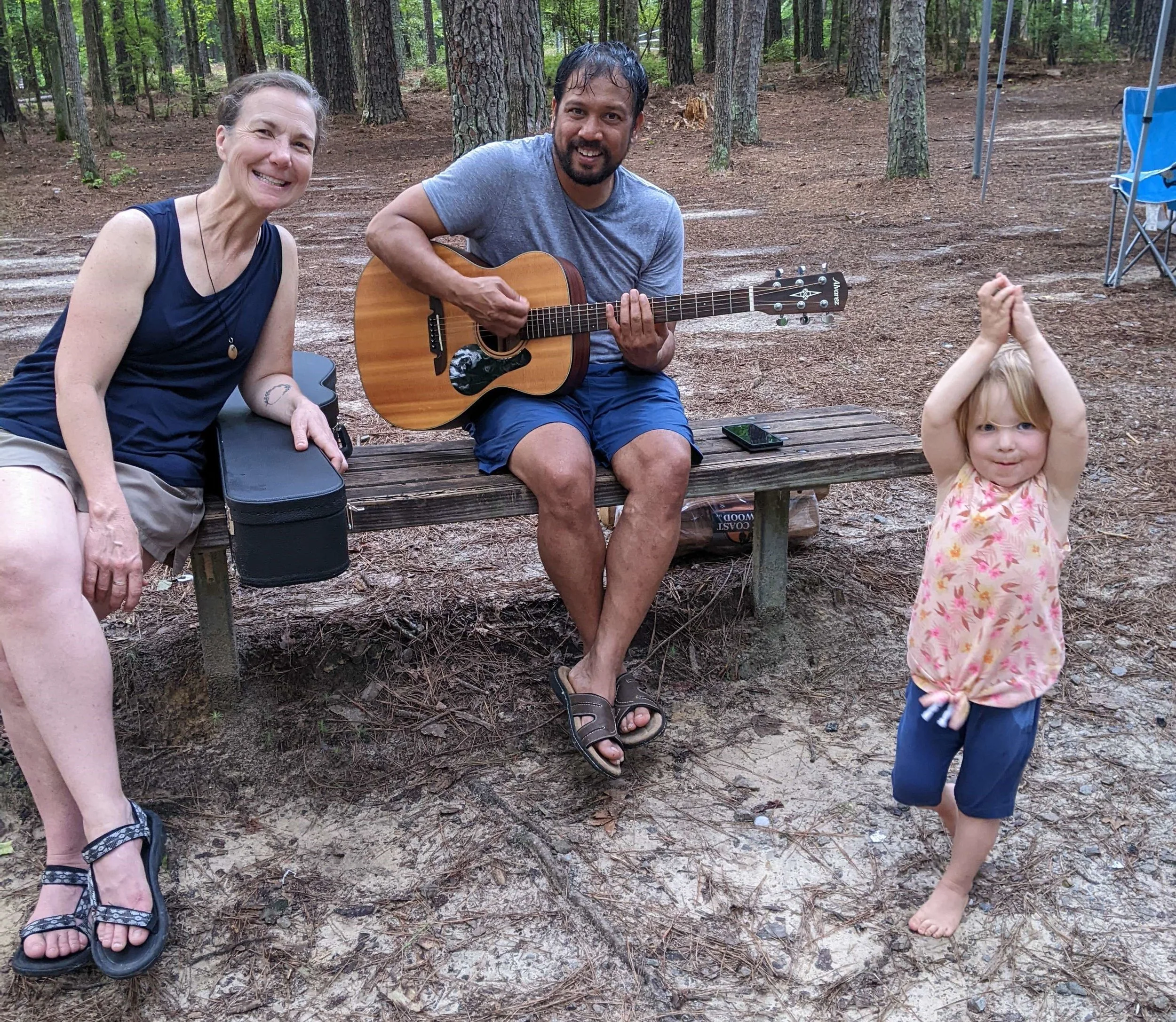 Two adults sitting on a bench outdoors, one playing a guitar, and a young child standing nearby in a playful pose in a wooded area.
