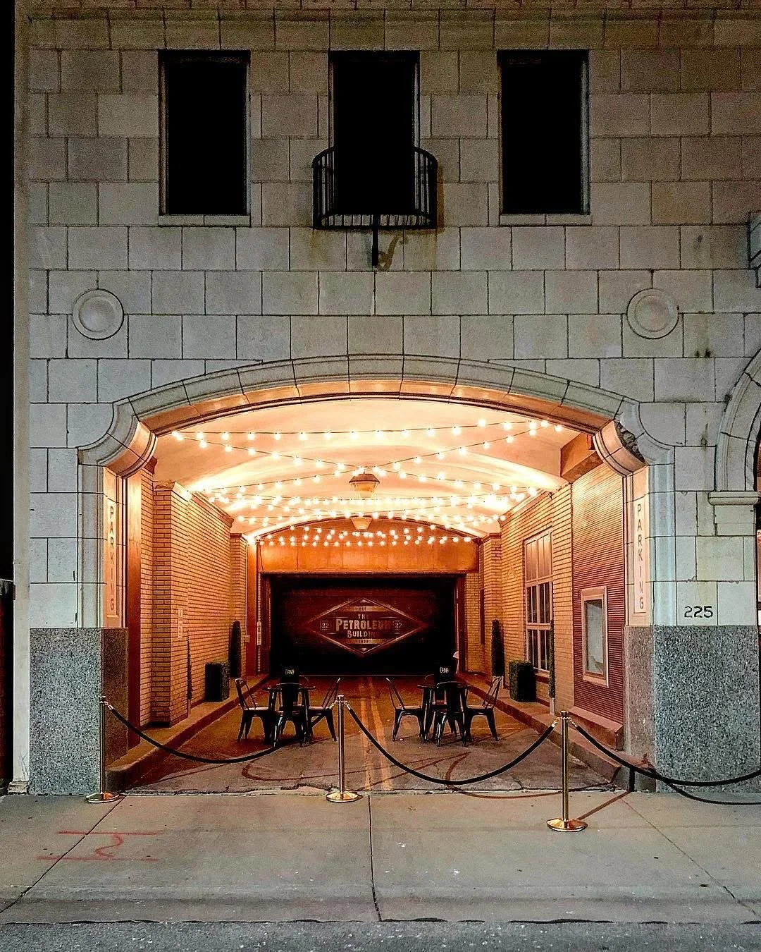 Night view of an open-air restaurant or bar area illuminated with string lights, with tables and chairs, enclosed by a roped barrier, adjacent to a building with a brick and stone facade.