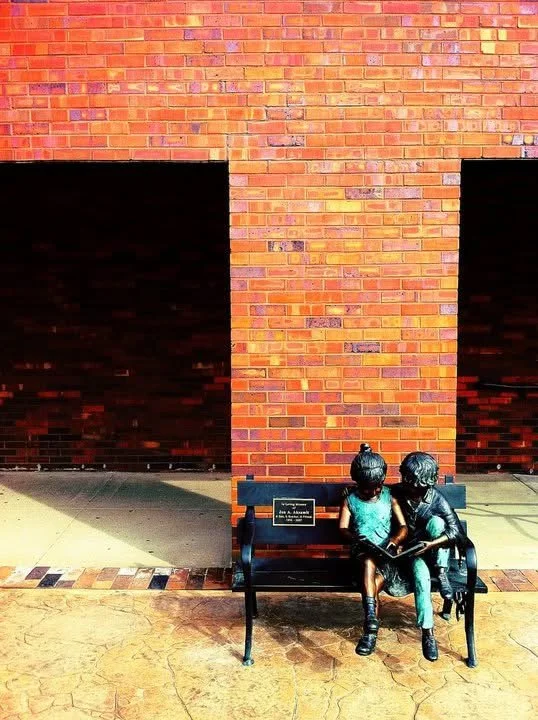Statue of two children sitting on a bench, with a brick wall in the background.