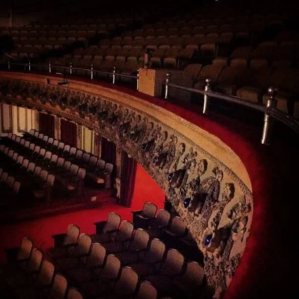 Interior view of a theater with rows of empty seats and a decorative balcony with painted artwork, dim lighting.