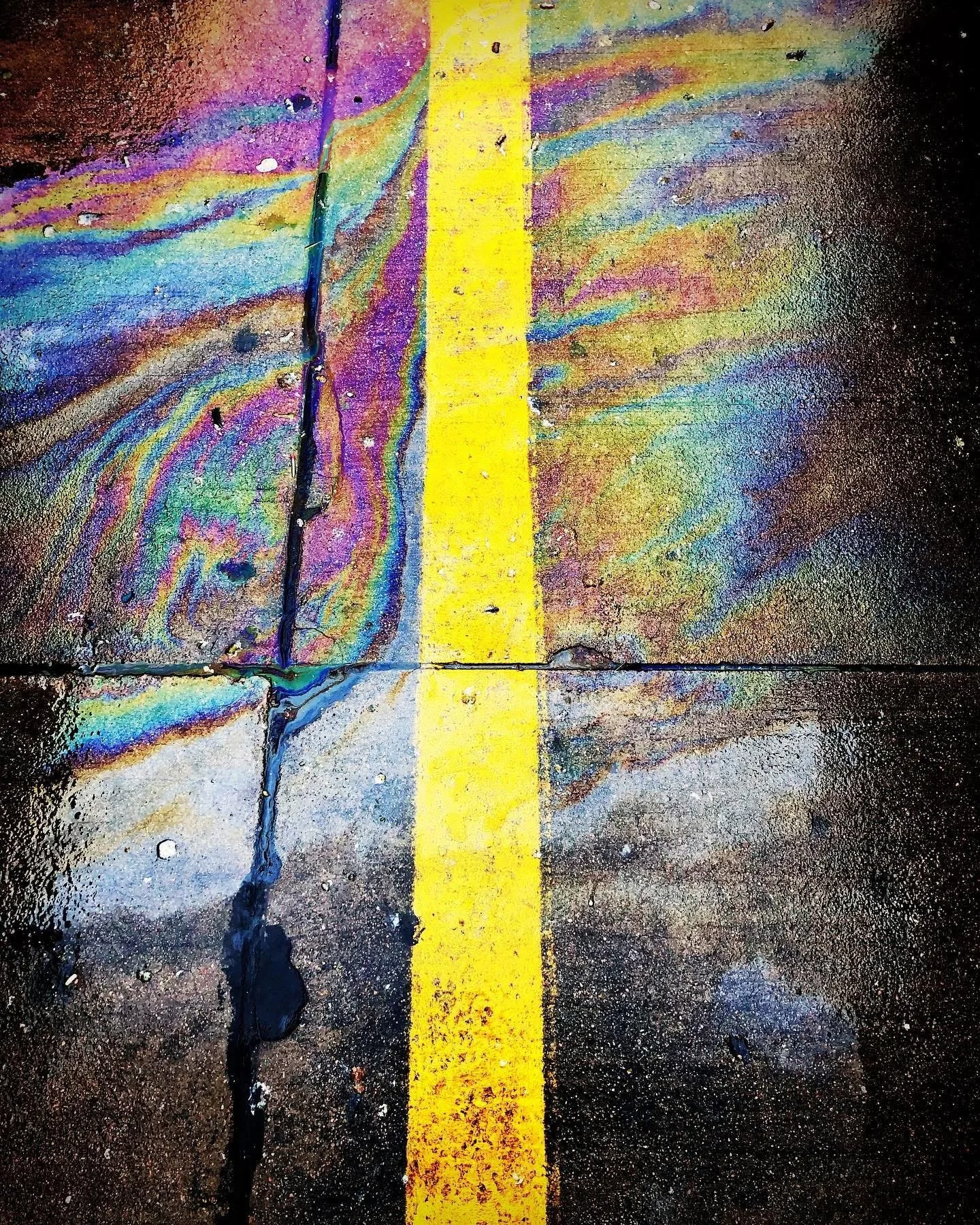 Wet pavement with a bright yellow line running vertically down the center and rainbow-colored oil slicks on both sides.