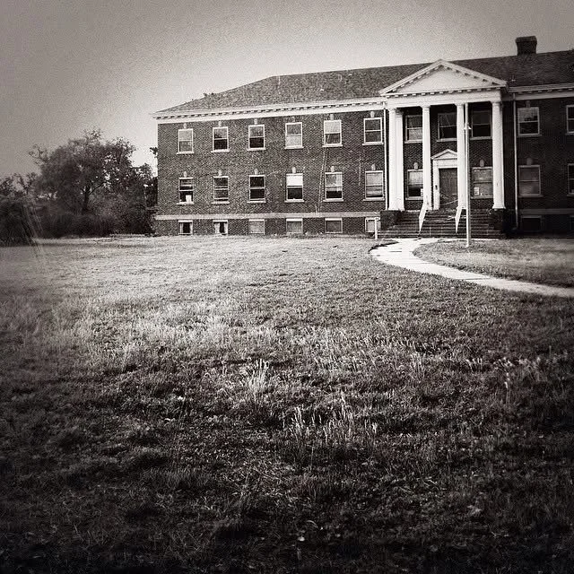 A large brick building with white columns and a gabled roof, set in a grassy area with a winding path leading to the entrance, in black and white.